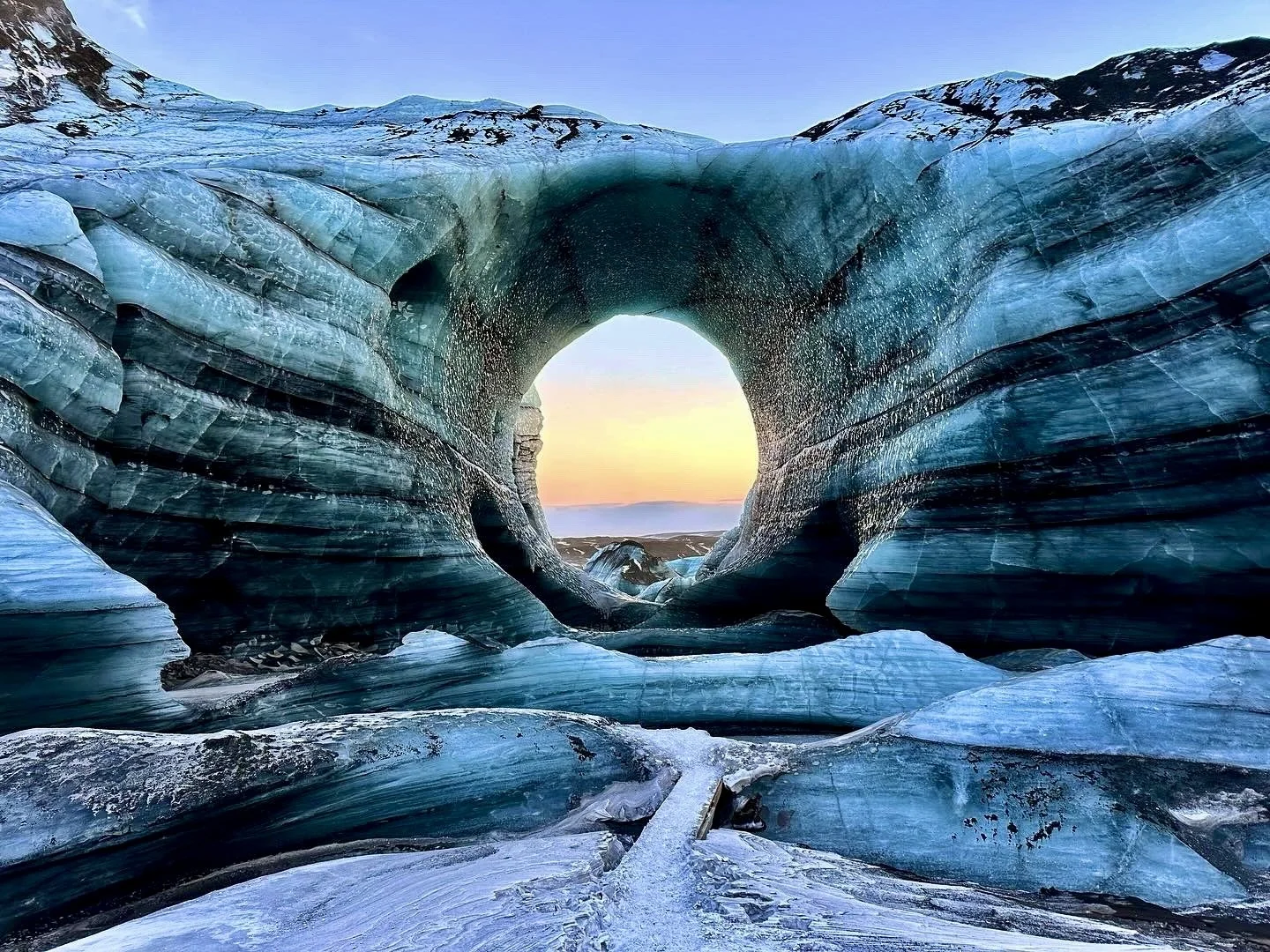 A circular opening in a wall of blue glacial ice frames a sunset sky at an ice cave site in Iceland.