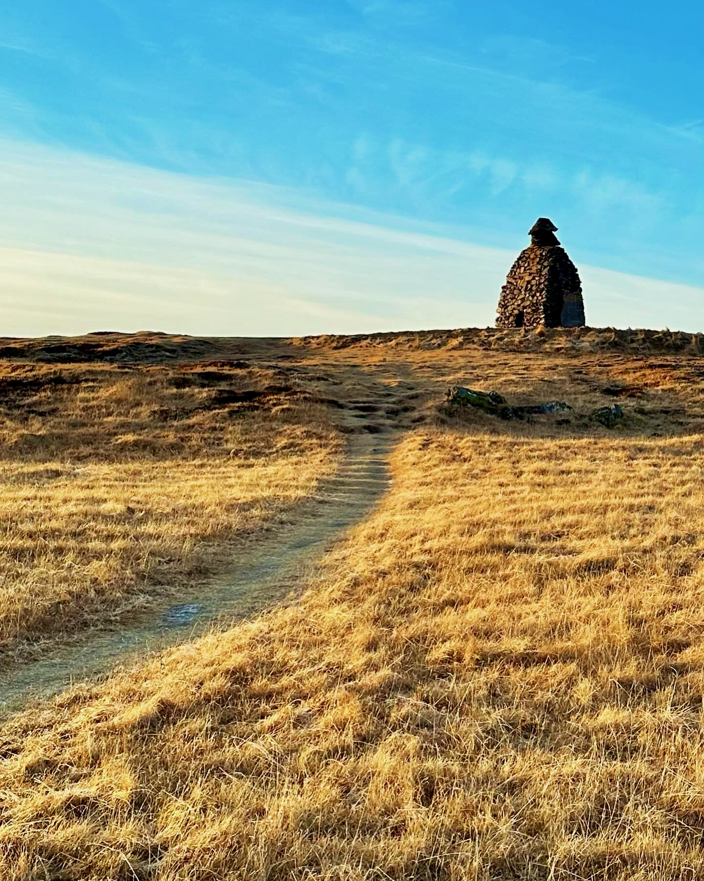 A dirt path winding through golden grassland toward a tall stone cairn on a hilltop in Iceland.