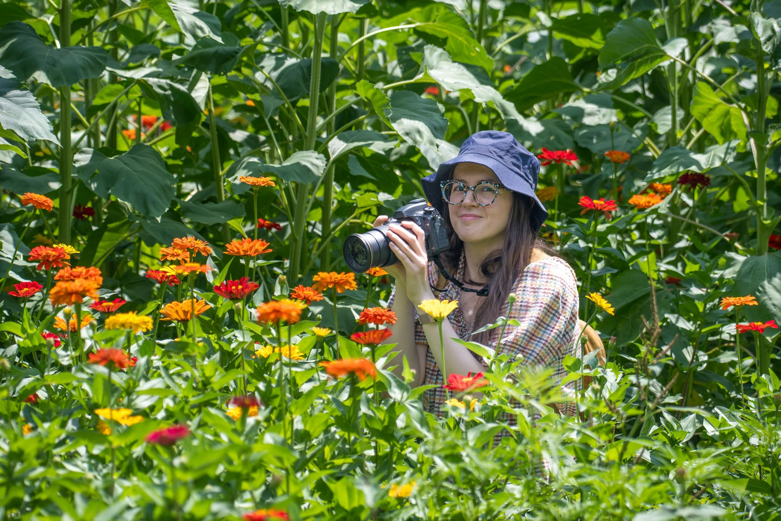 Kristin holding a camera while standing among tall green plants and orange, red, and yellow flowers.