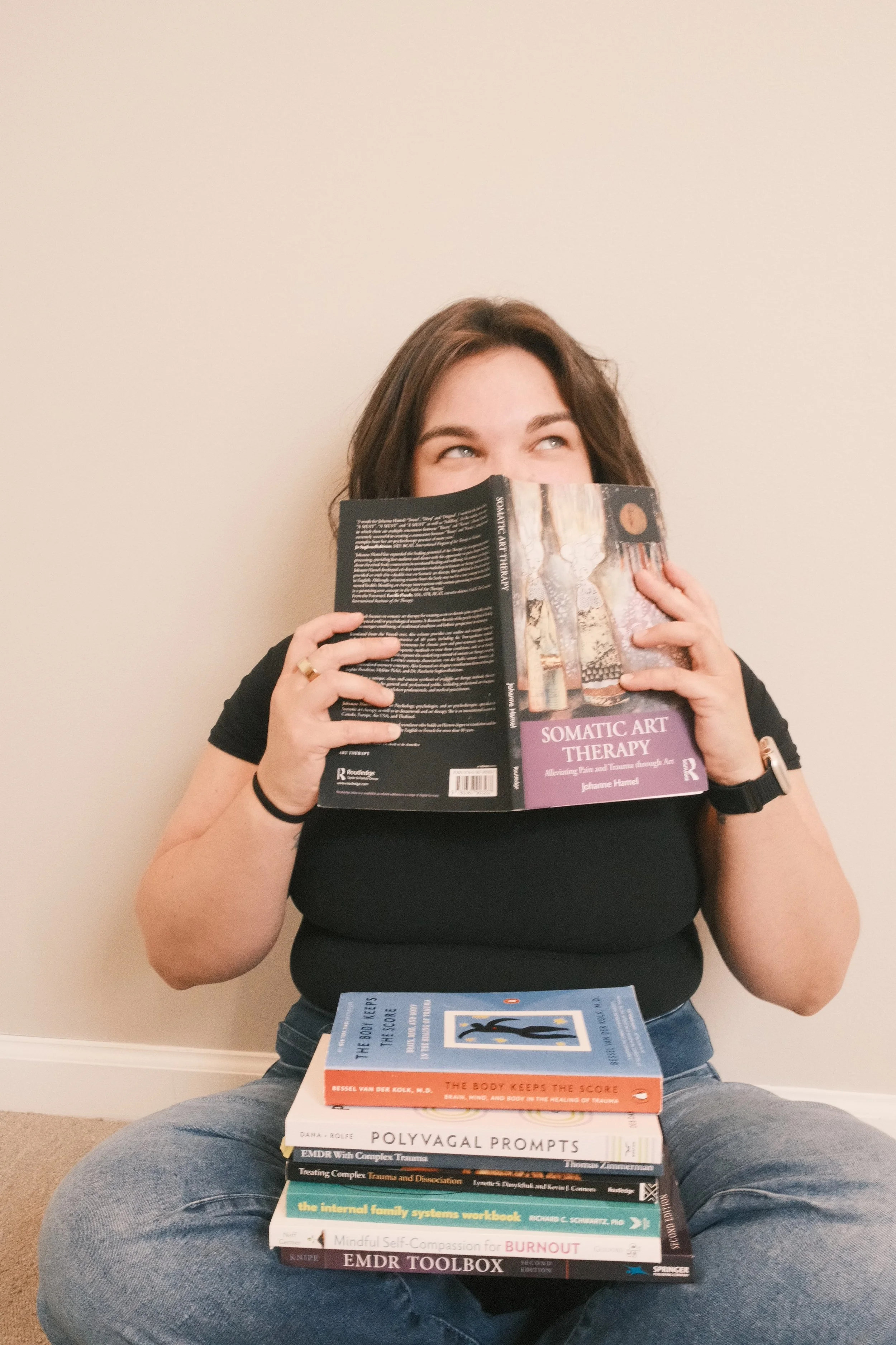 A woman with brown hair sitting cross-legged against a beige wall, reading a book titled 'Somatic Art Therapy.' There is a stack of books on her lap.