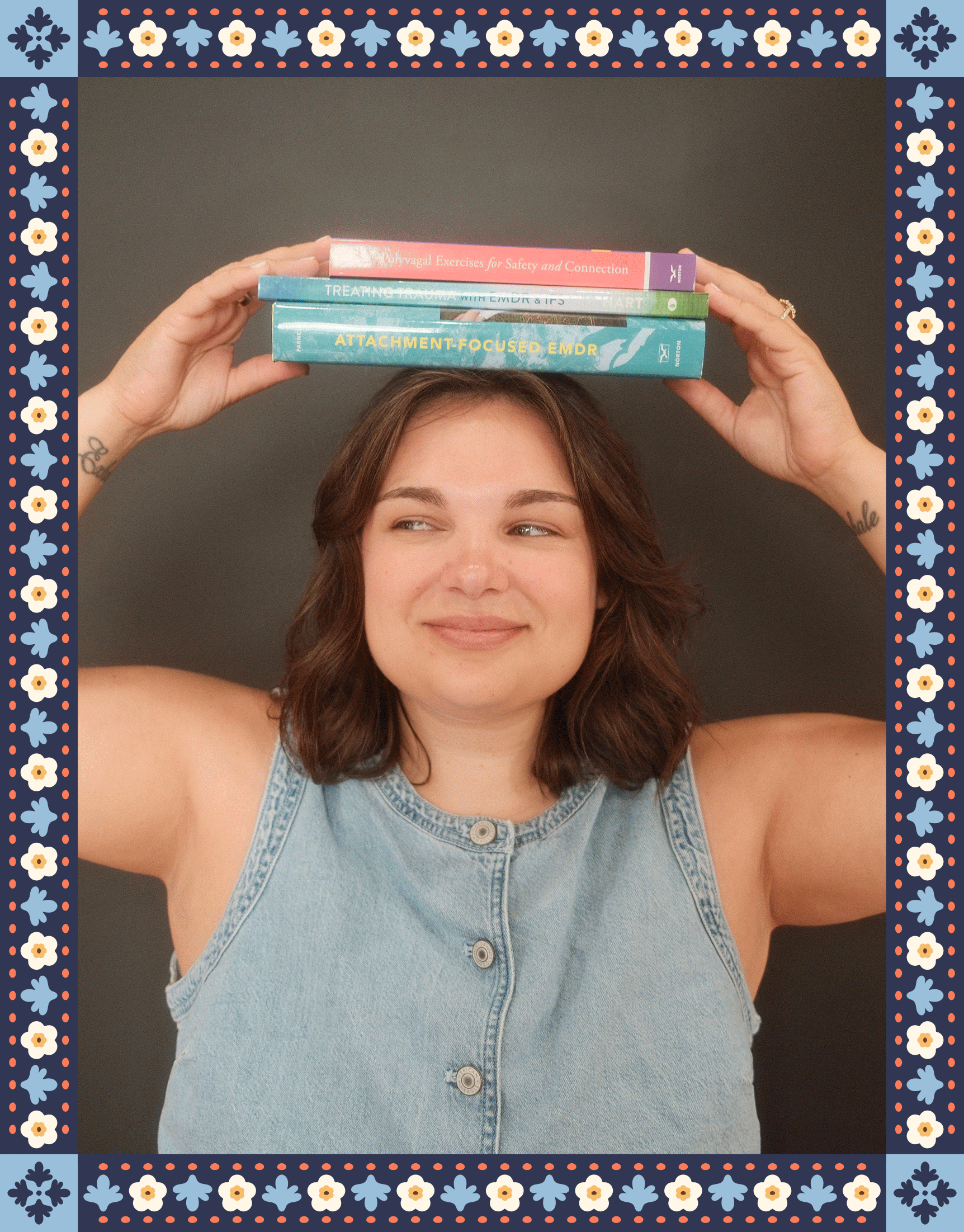 A woman with shoulder-length brown hair is holding three books above her head. She is smiling and wearing a sleeveless denim top. Two tattoos can be seen on her arms, and she is against a plain background with a decorative border featuring snowflakes and flowers.