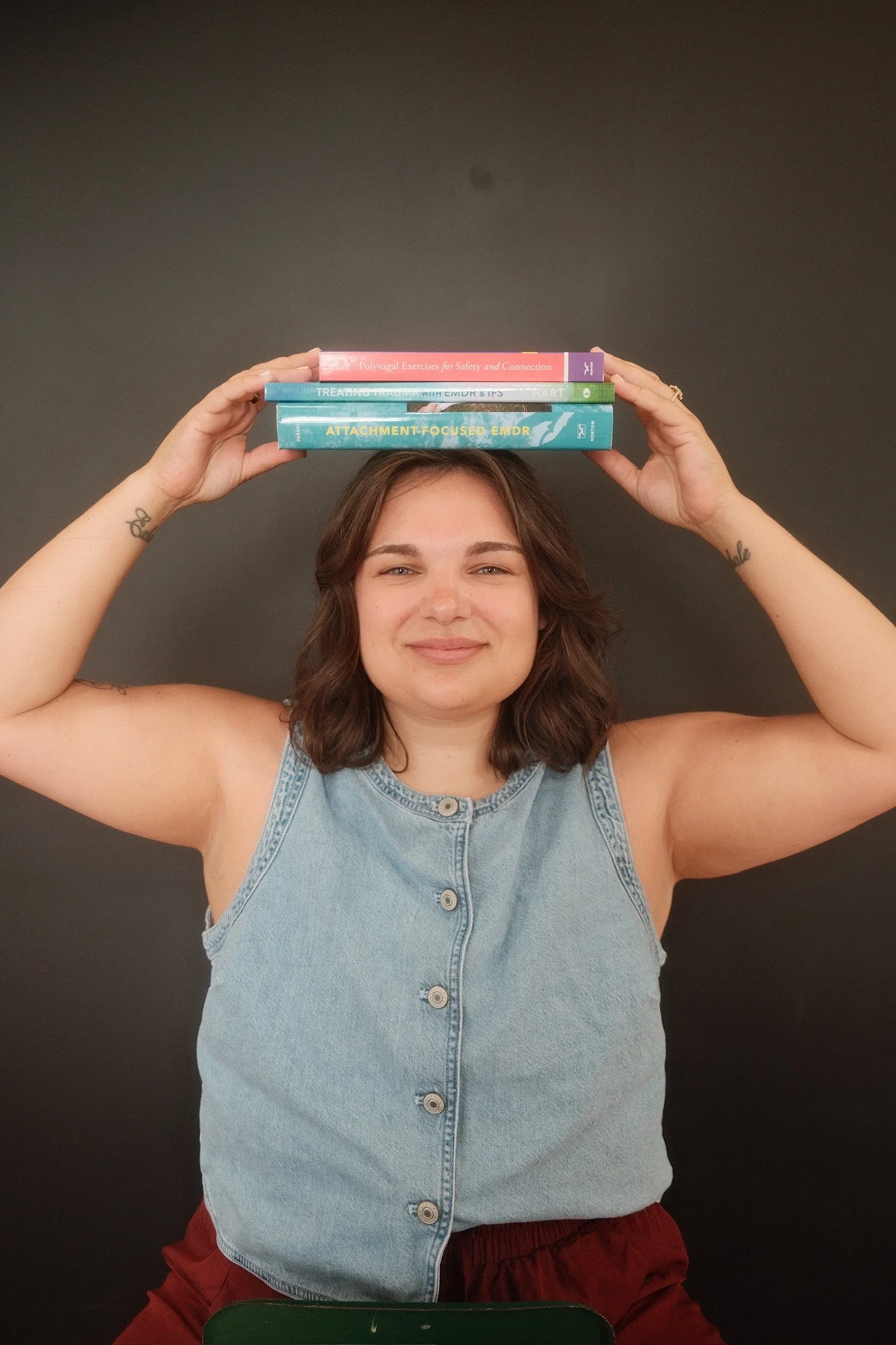 A woman with medium-length brown hair, wearing a sleeveless denim top and red shorts, balancing four books on her head, smiling, against a dark background.