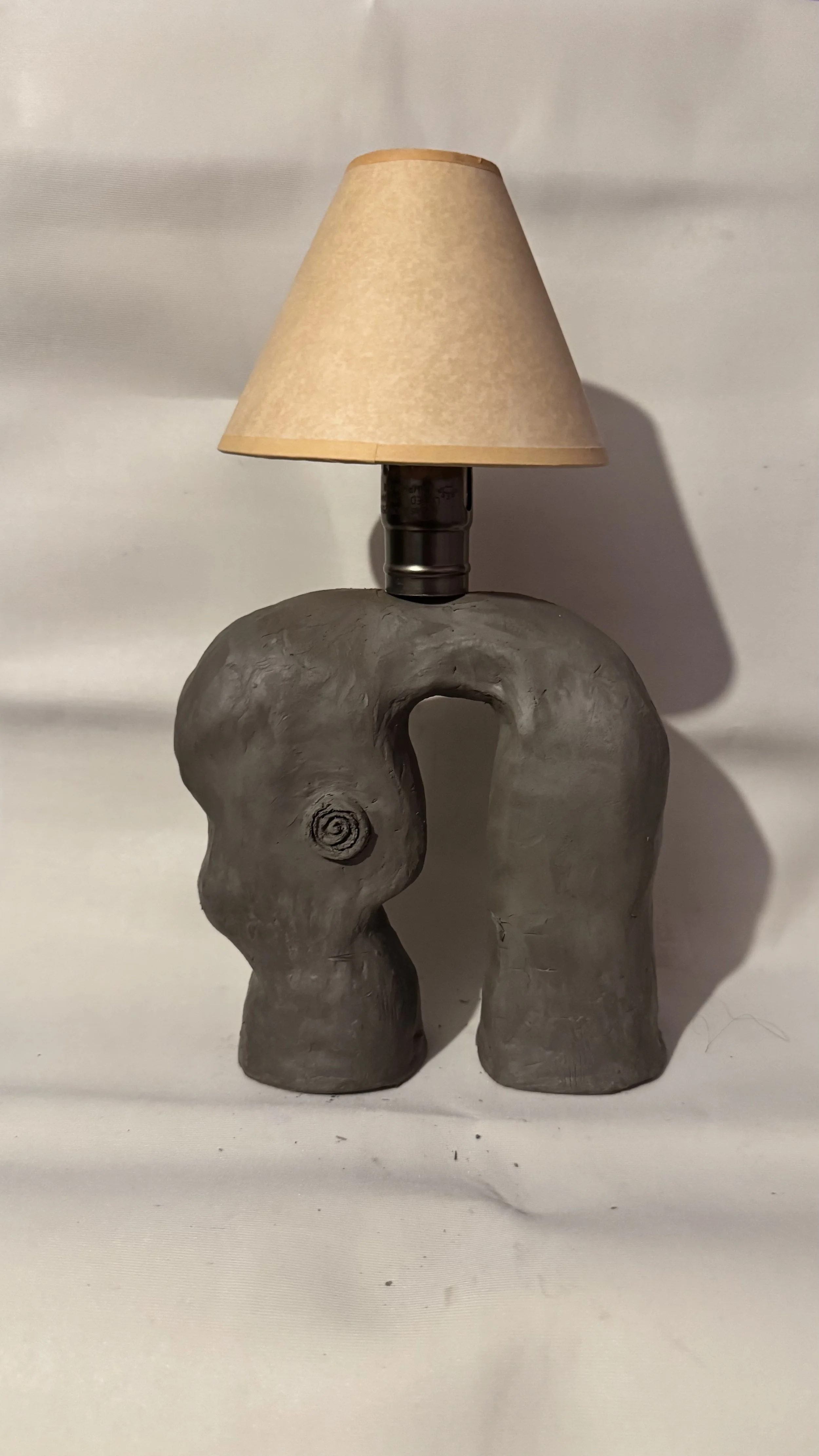 A table lamp with a base shaped like a gray, sculpted elephant head with a curled trunk and a circular eye, topped with a beige lampshade.