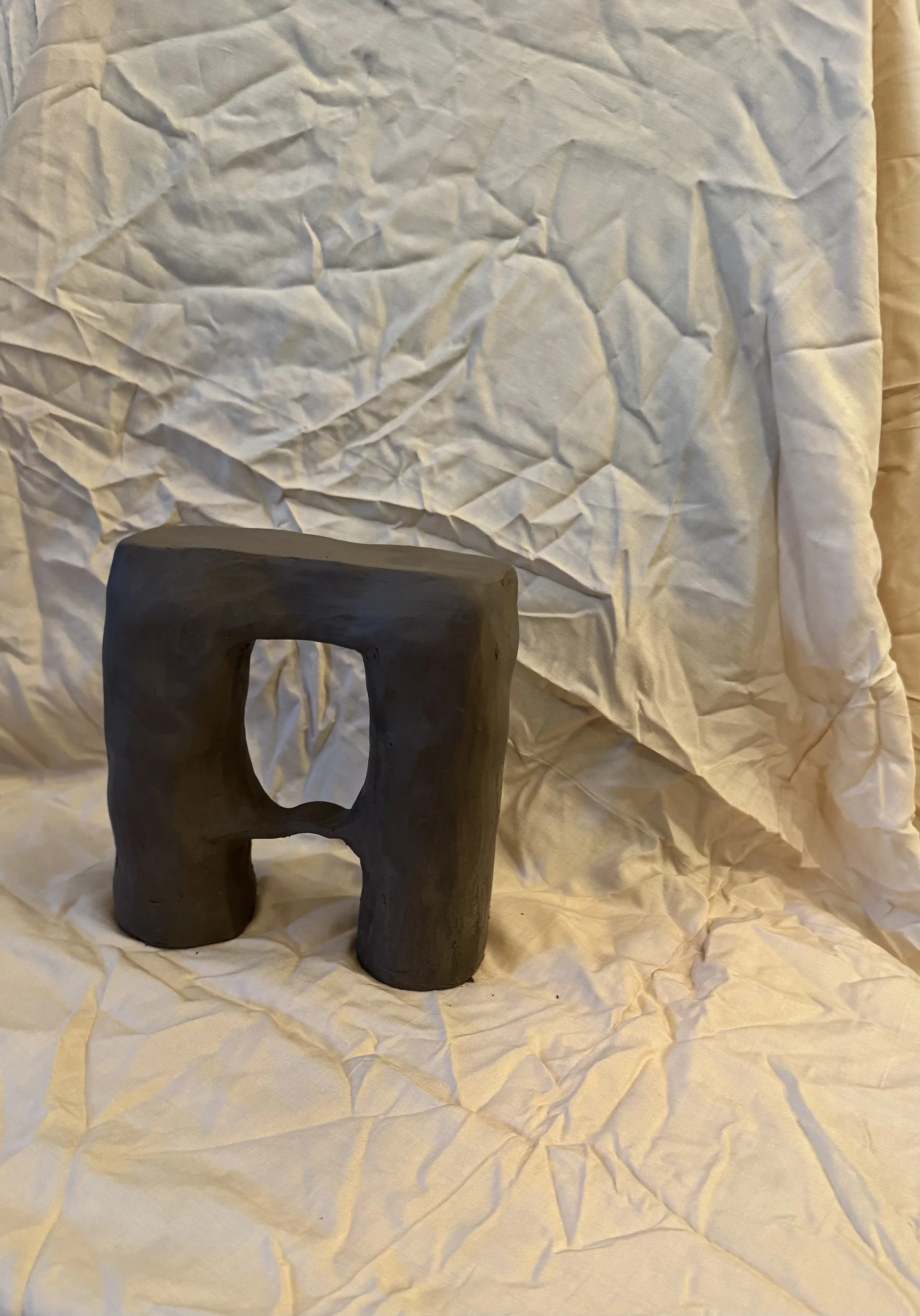 A small, abstract sculpture made of dark clay on crumpled white paper, with a hollow section in the middle, setting against more crumpled white paper backdrop.