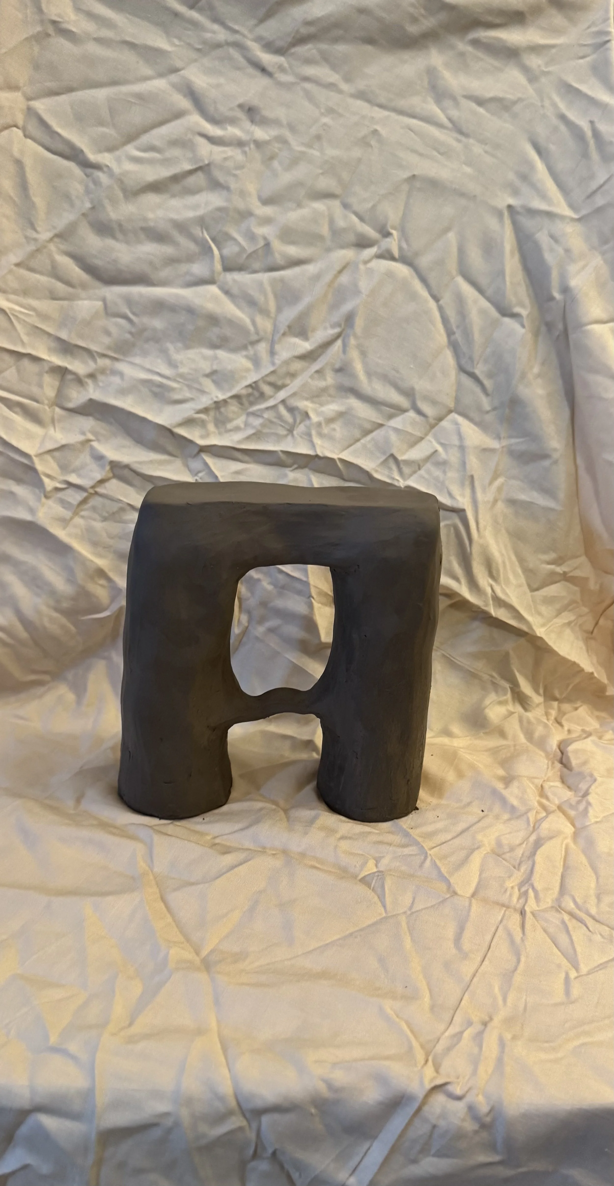 A small carved sculpture resembling the letter 'A' with a hole in the middle, made of dark material, placed on crumpled beige paper with a crumpled background.