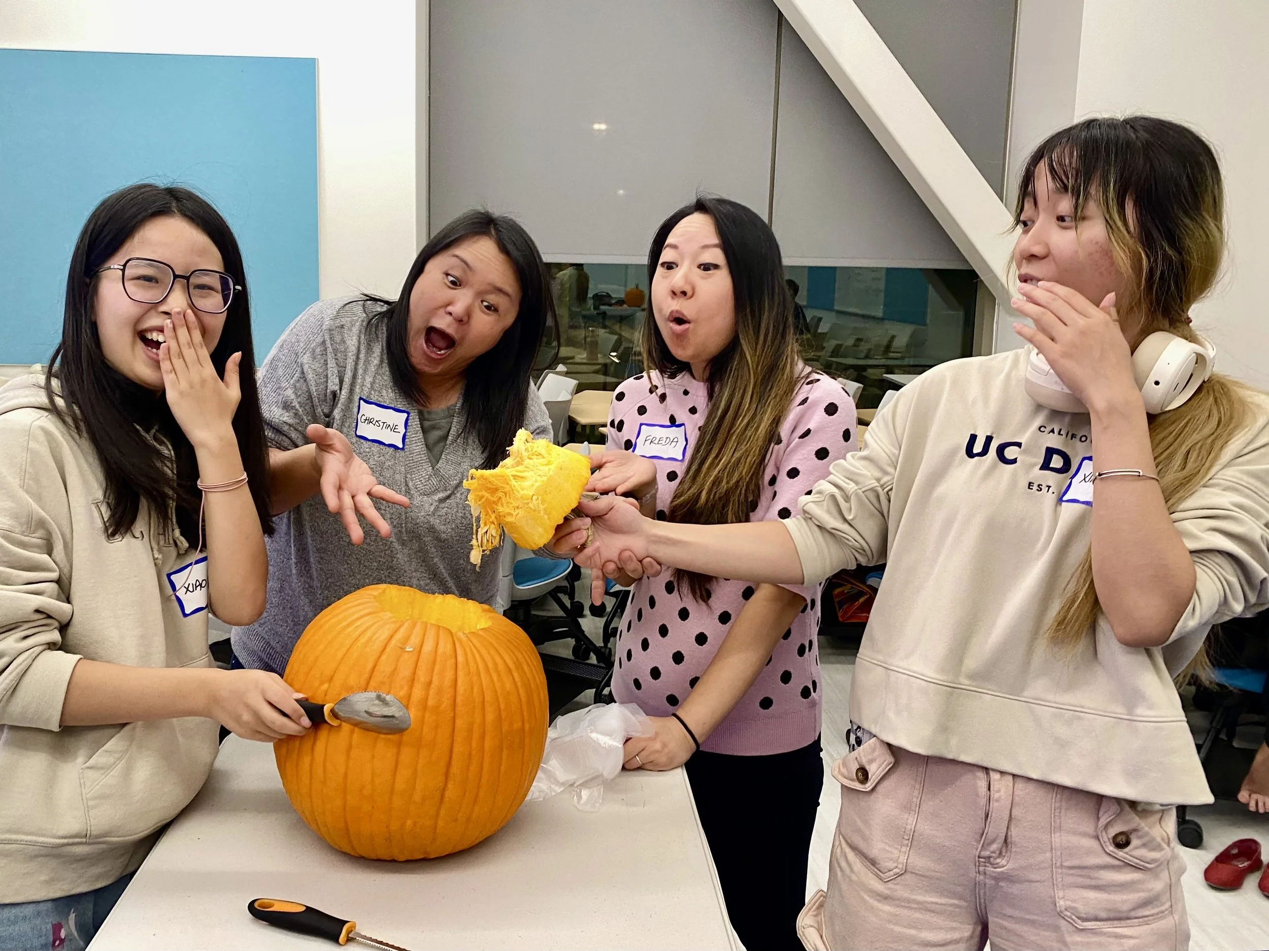 Four women standing around a table with a large pumpkin, one woman is scooping out the pumpkin, all appear surprised or amused, in a room with large windows and blue wall panels, wearing casual clothing and name tags.