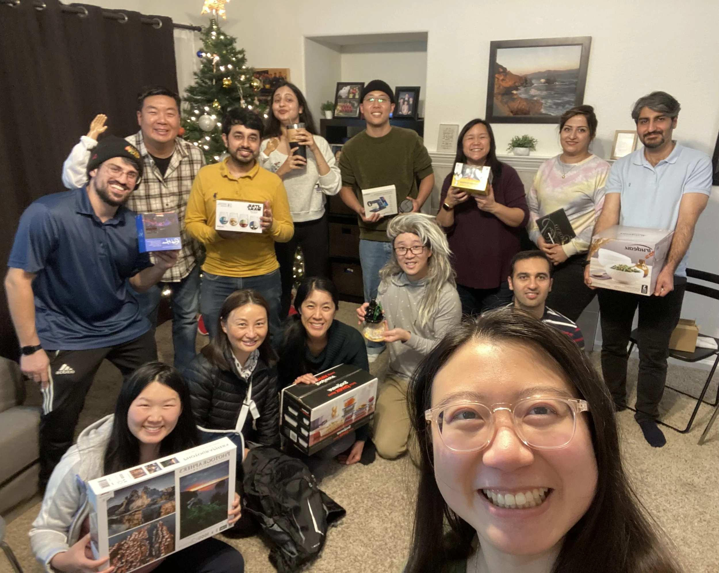 A group of 15 people celebrating in a living room with a decorated Christmas tree, holding various board games and gifts, smiling for a group photo.