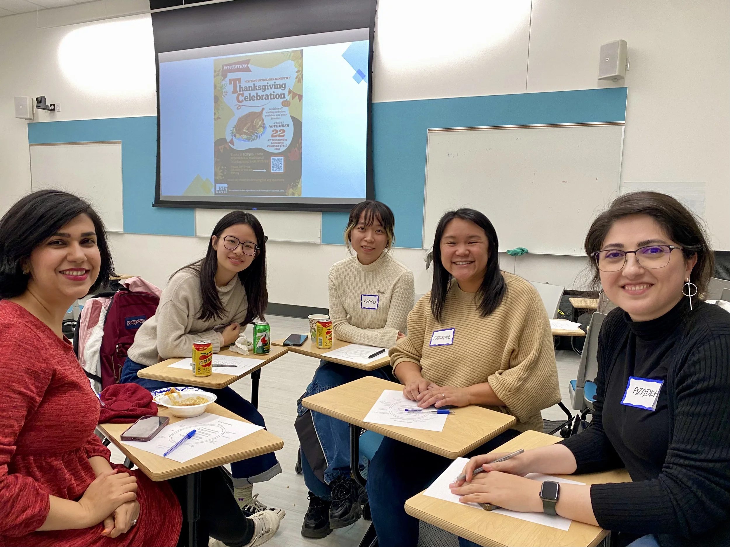Group of five women sitting at desks with notebooks, pens, and drinks, smiling at the camera in a classroom. A presentation slide with a Thanksgiving celebration poster is projected on a screen in the background.
