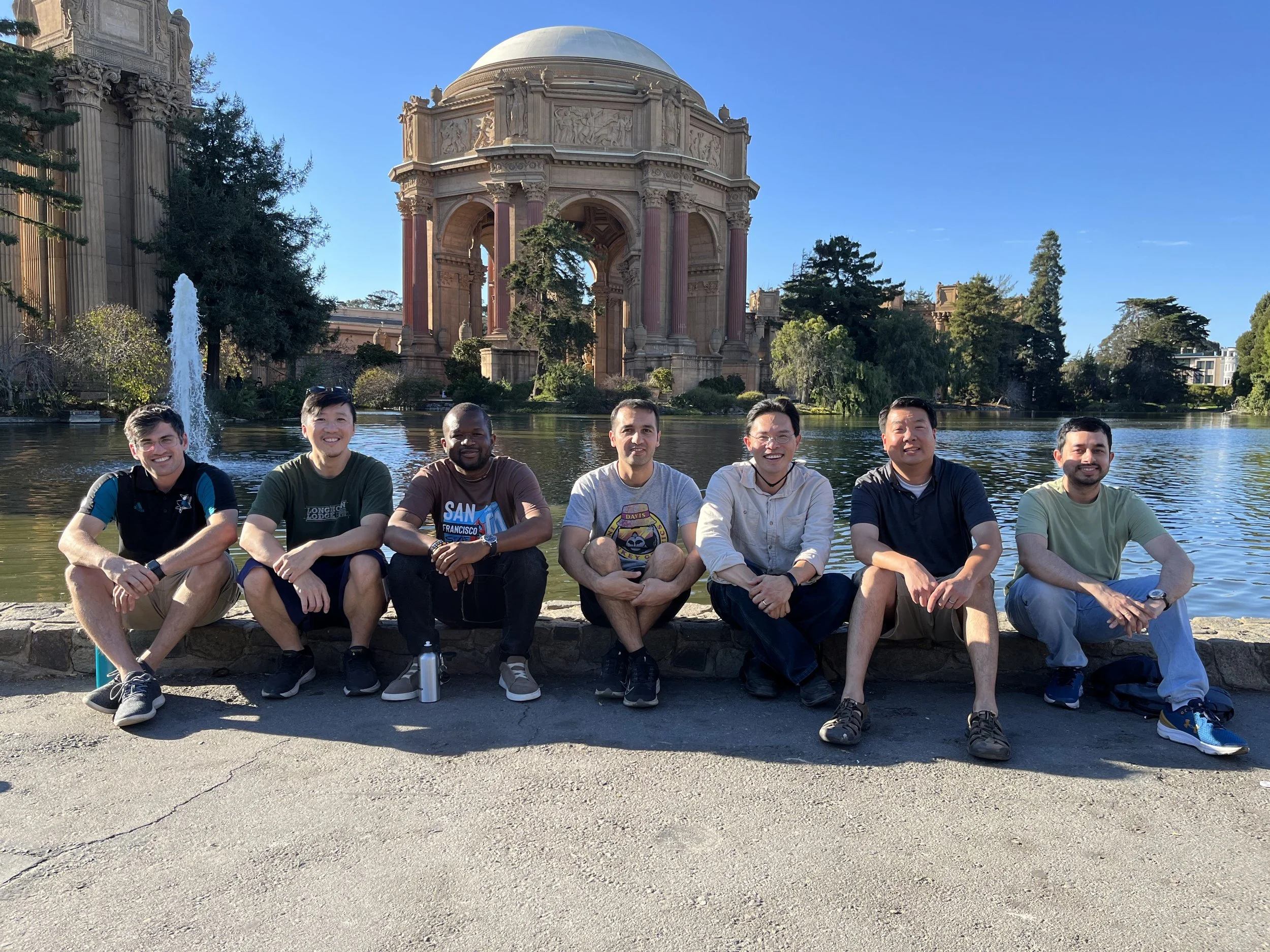 Seven men sitting on a stone ledge in front of a lake, with a large classical building and trees in the background, during the daytime.