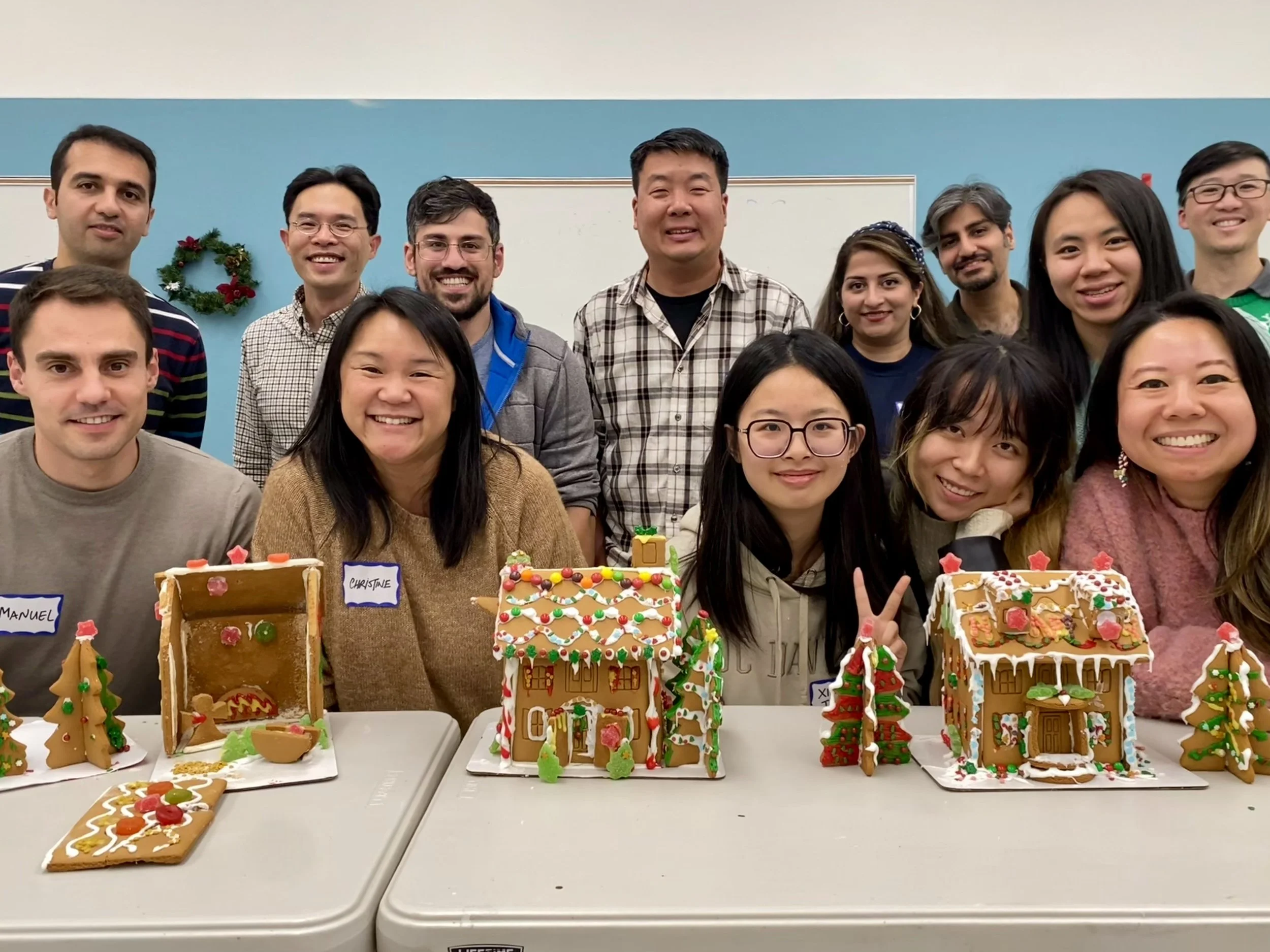 Group of people celebrating Christmas with decorated gingerbread houses and cookies, smiling in a festive indoor setting.