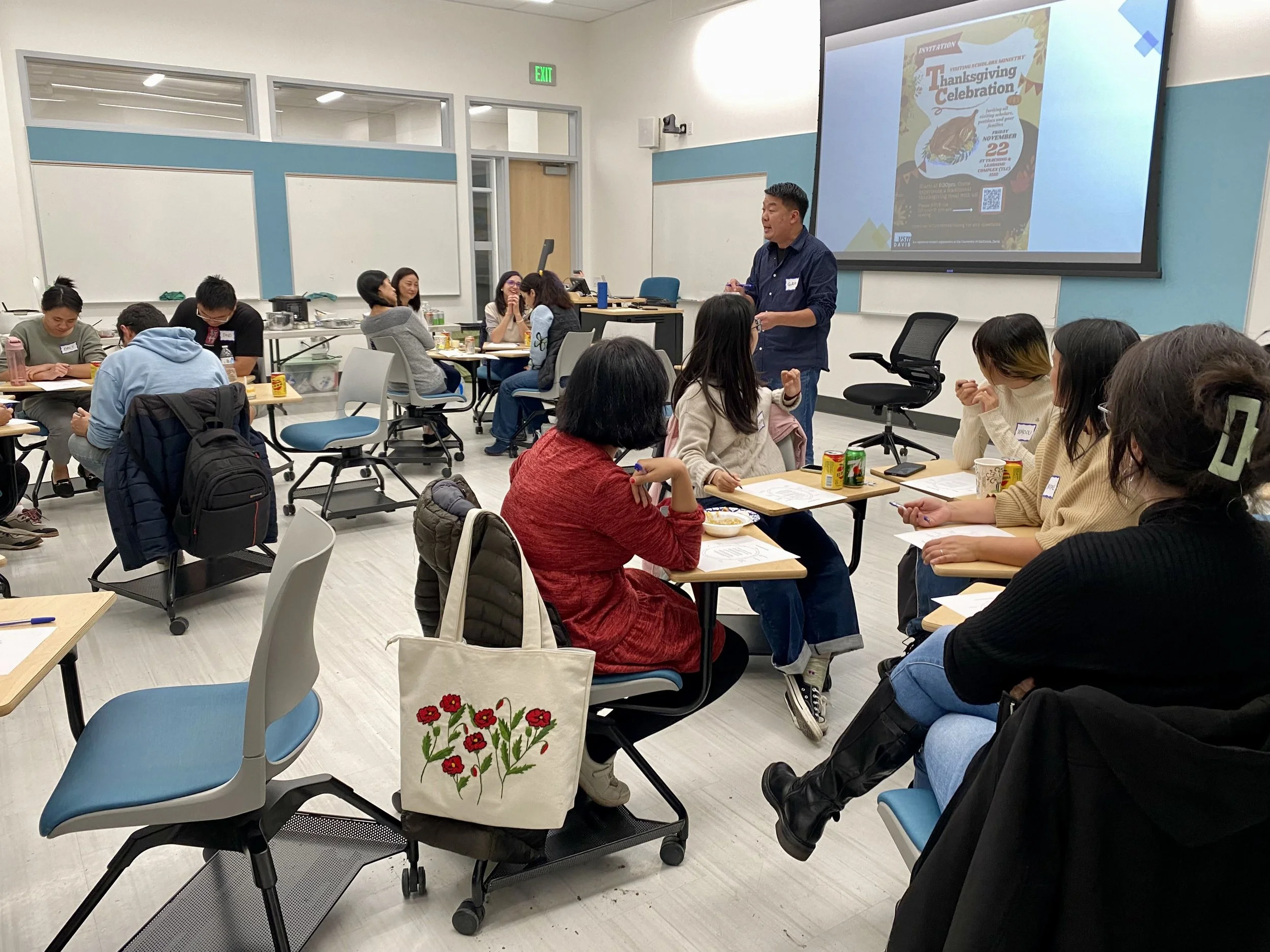 A classroom or conference room with students sitting at tables, listening to a speaker at the front. The speaker is standing near a large screen displaying a poster for a Thanksgiving celebration. There are notebooks, drinks, and snacks on the tables