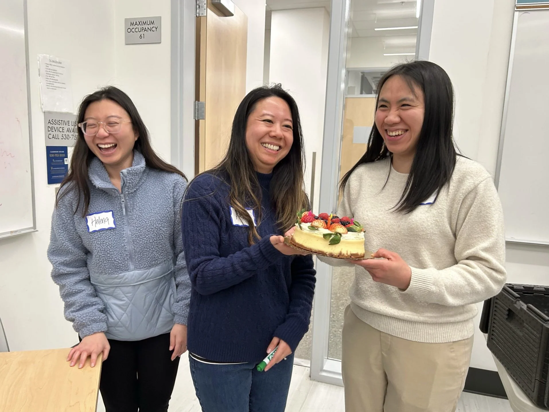 Three women celebrating, one of whom is holding a fruit-topped birthday cake. They are indoors, smiling and laughing.