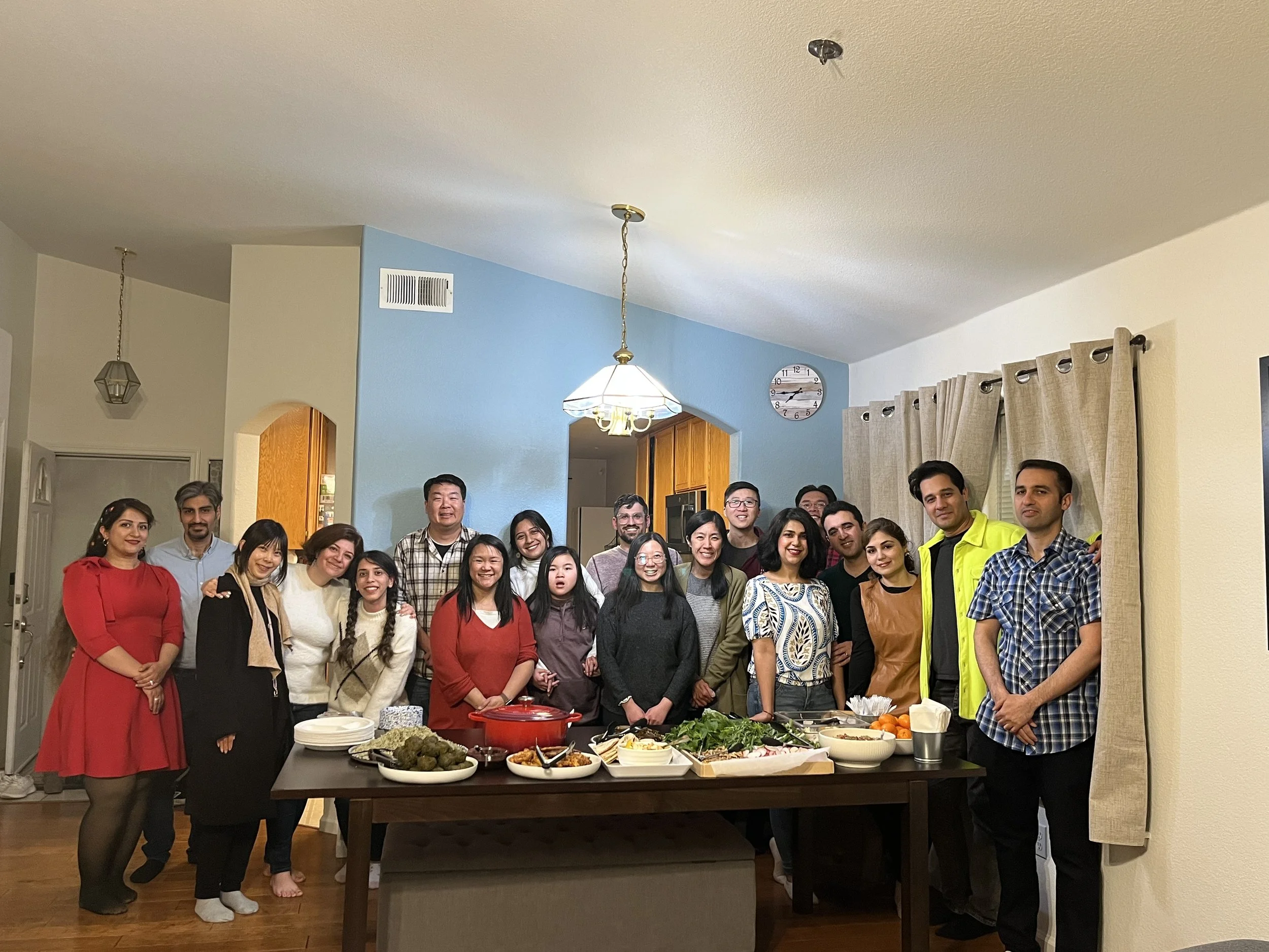 A group of approximately 20 people gathered in a dining room, standing behind a table with food, celebrating together. The room has a blue accent wall, beige curtains, and a clock on the wall. Some people are smiling and dressed casually or semi-form