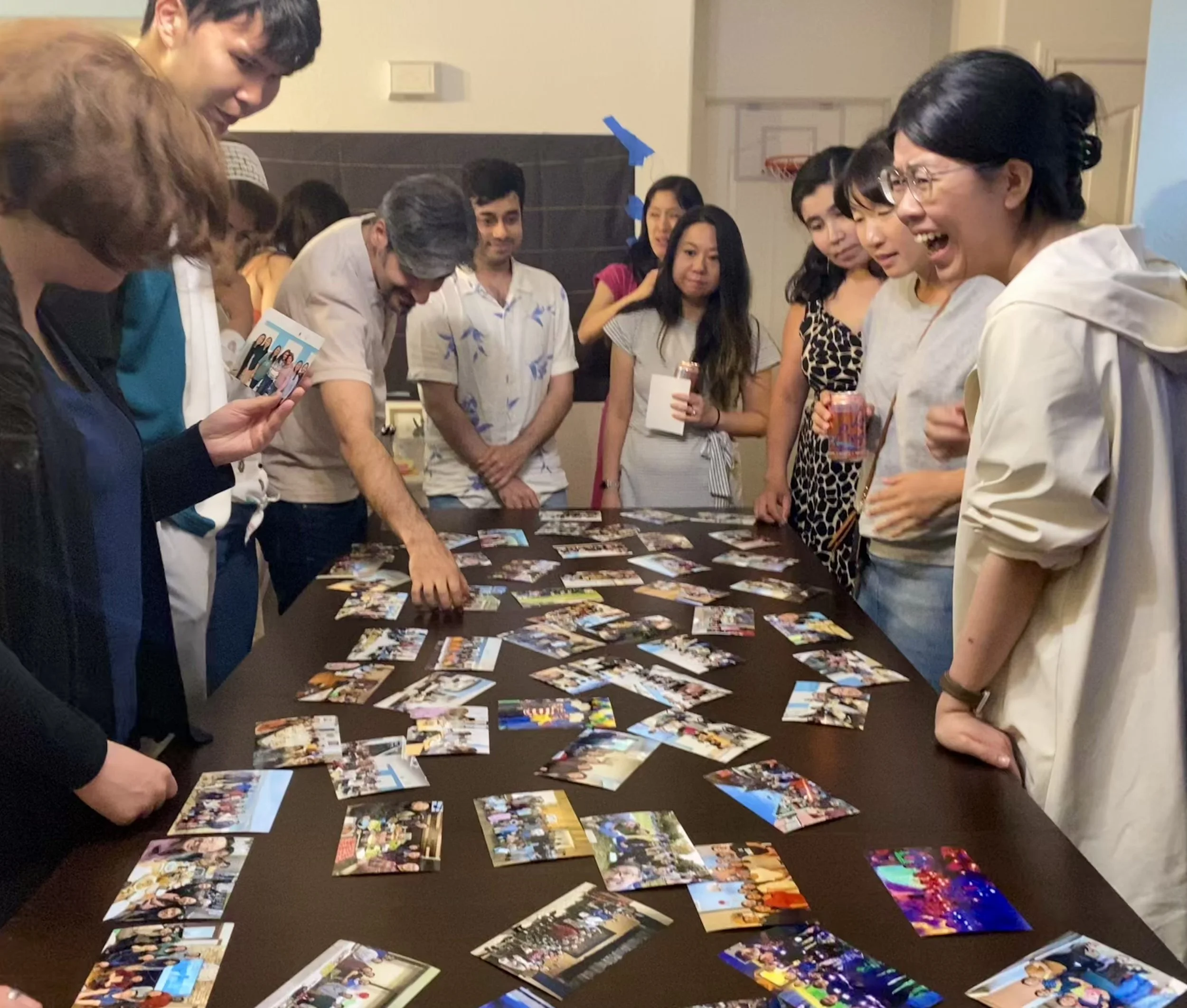 Group of people gathered around a table looking at and arranging printed photographs, at a social event or gathering.