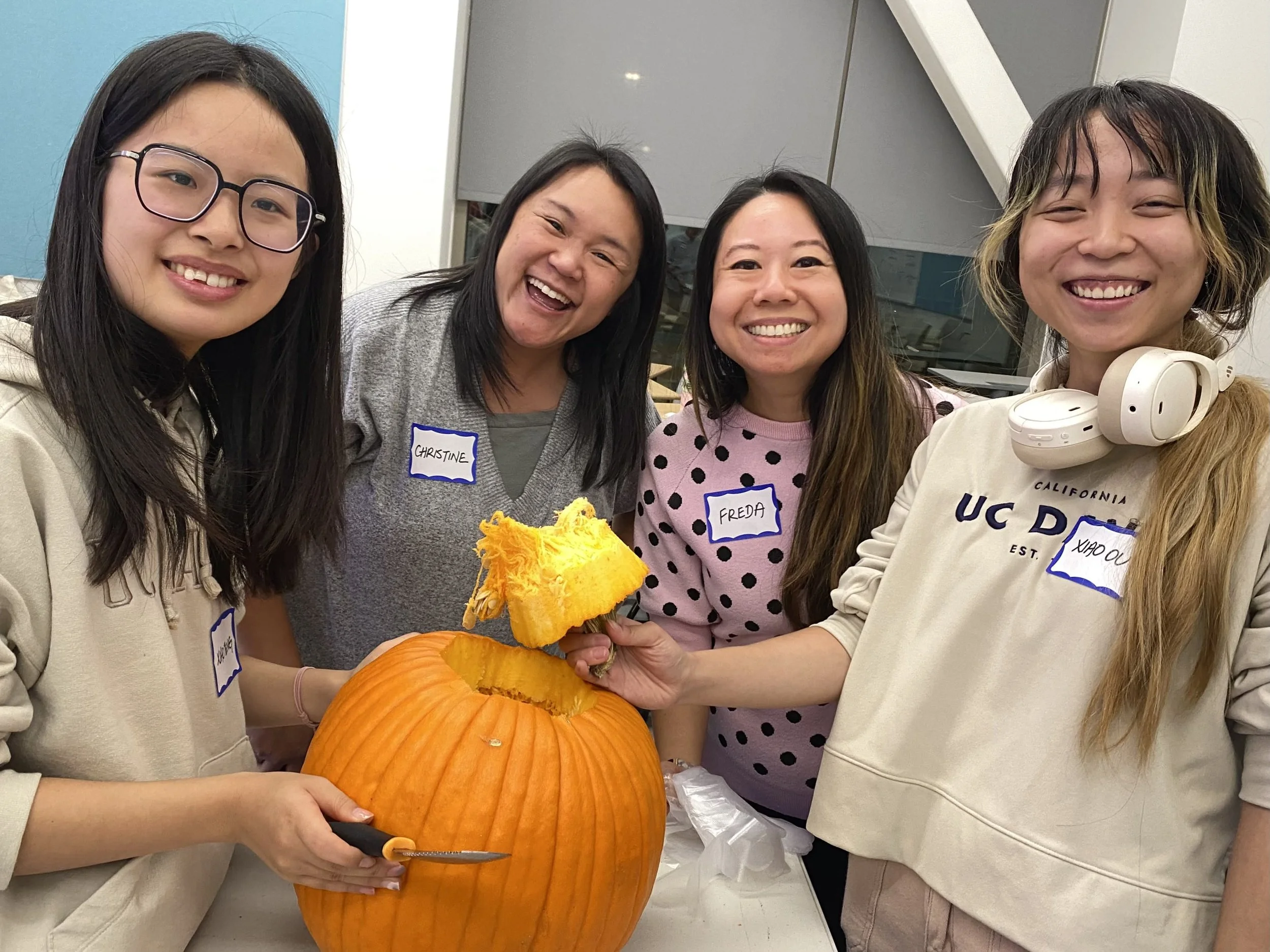 Four women smiling and gathered around a large pumpkin, with one holding a piece of pumpkin that they are about to carve.