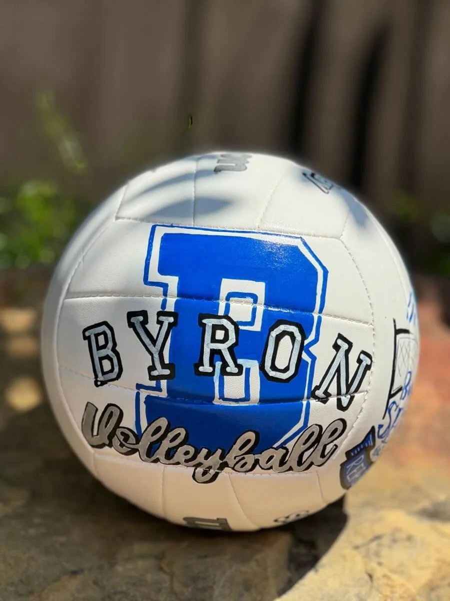 White volleyball featuring a big, blue "B" and "Byron volleyball" written over top in grey, sitting on a large rock.