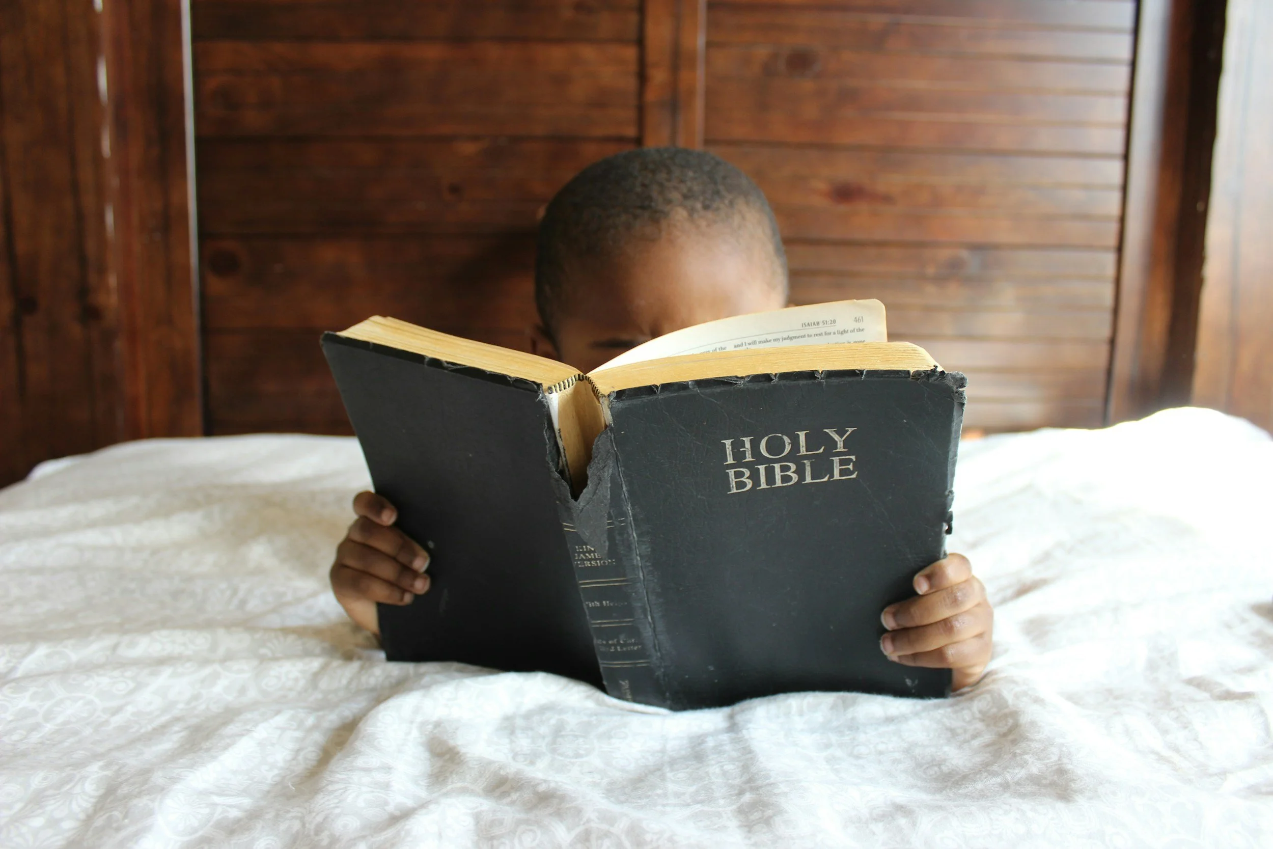 A child sitting on a bed, reading a black "Holy Bible."
