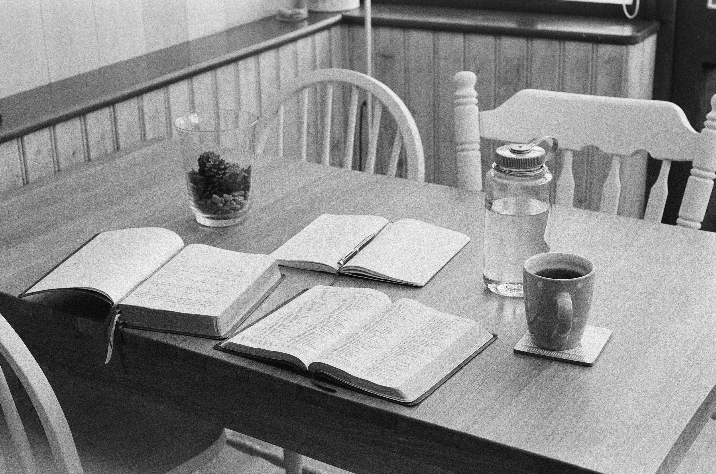An open Bible and custom prayer journal on a wooden table, with a plant and water bottle beside them.