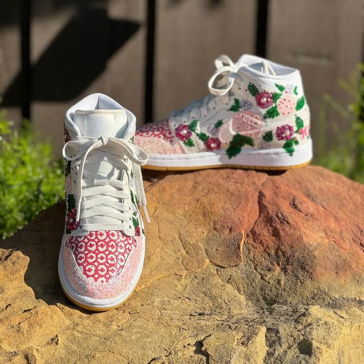 A pair of white high-top sneakers decorated with pink and green floral patterns, placed on a large rock outdoors.