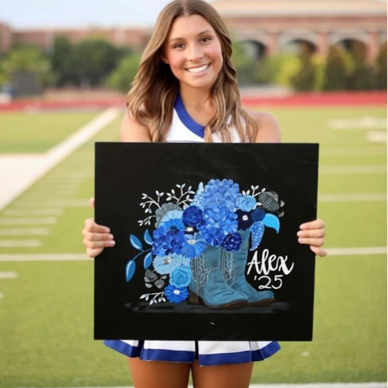 Cheerleader smiling while holding a custom cheer box board of a boots and flowers scene on a football field.