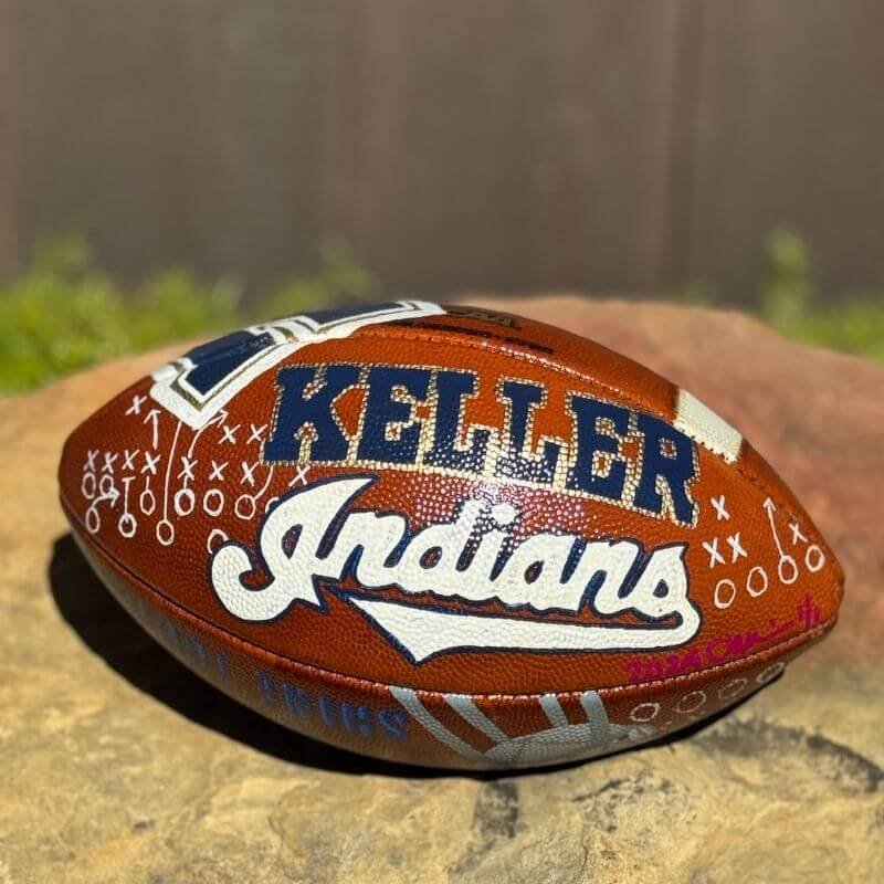 A football with "Keller Indians" and football play markings, placed on a rock surface outdoors.