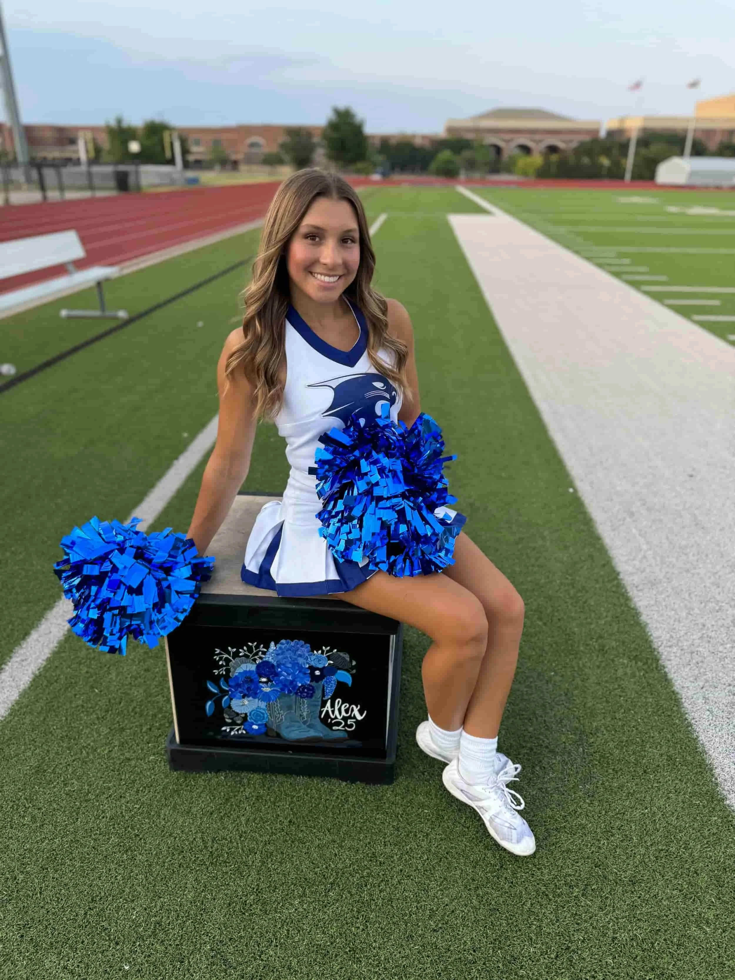 Young female cheerleader in a white and blue uniform sitting on a box with a floral design and the name 'Alex 25' written on it, holding blue pom-poms, on a football field at dusk.