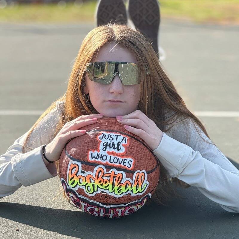Girl with long hair wearing sunglasses, lying on a basketball court holding a basketball with text "Just a girl who loves basketball."