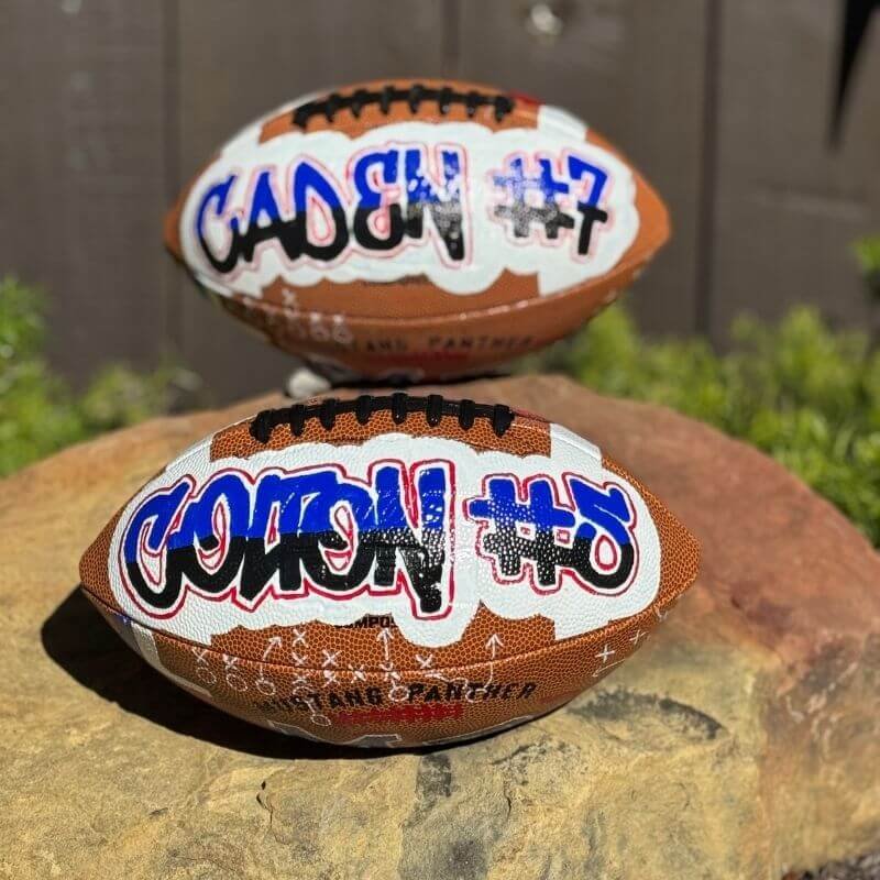 Close-up of two American footballs with graffiti-style designs and player numbers, resting on a rock outdoors.