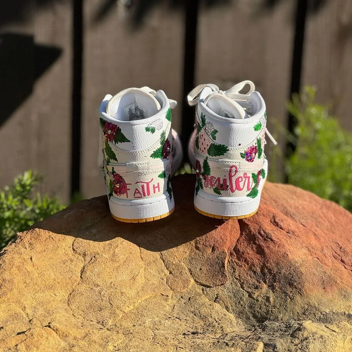 A pair of white high-top sneakers decorated with floral and berry patterns, with the words 'FAITH' and 'FLOUR' written on the back, placed on a large rock outdoors.