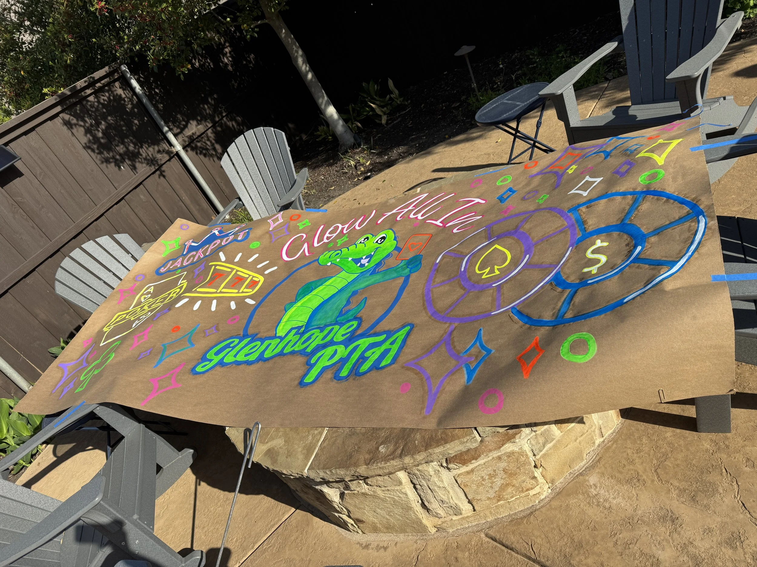 Colorful, hand-painted banner on a table with various illustrations, including an alligator, poker chips, dollar signs, and the words 'Glow All In' and 'Glenhope PTA' written in bright colors.