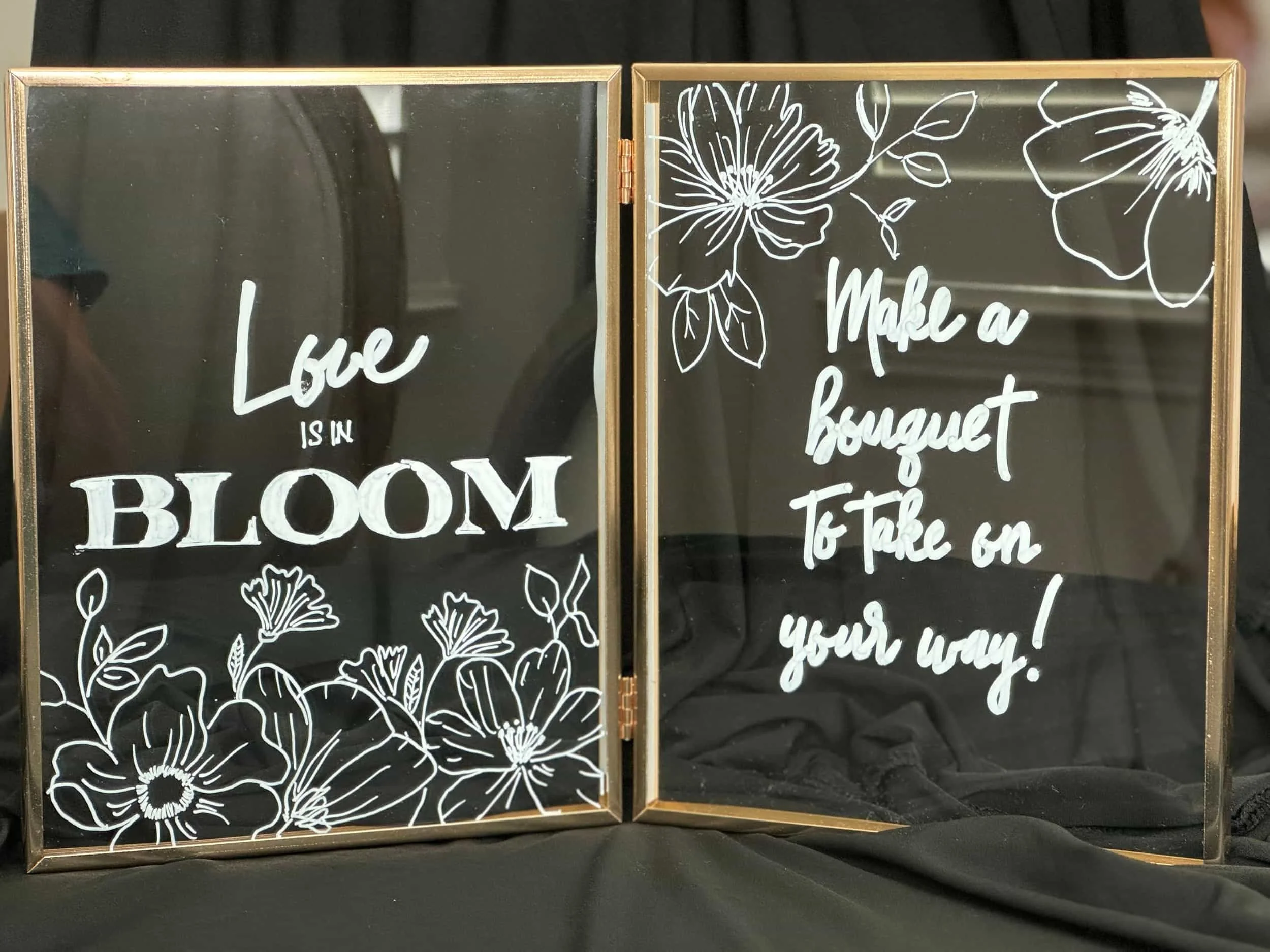 Decorative black-framed mirrors with white floral drawings and inspirational quotes. One reads 'Love is in bloom' with flowers, the other says 'Make a bouquet to take on your way!' with flowers at the top.