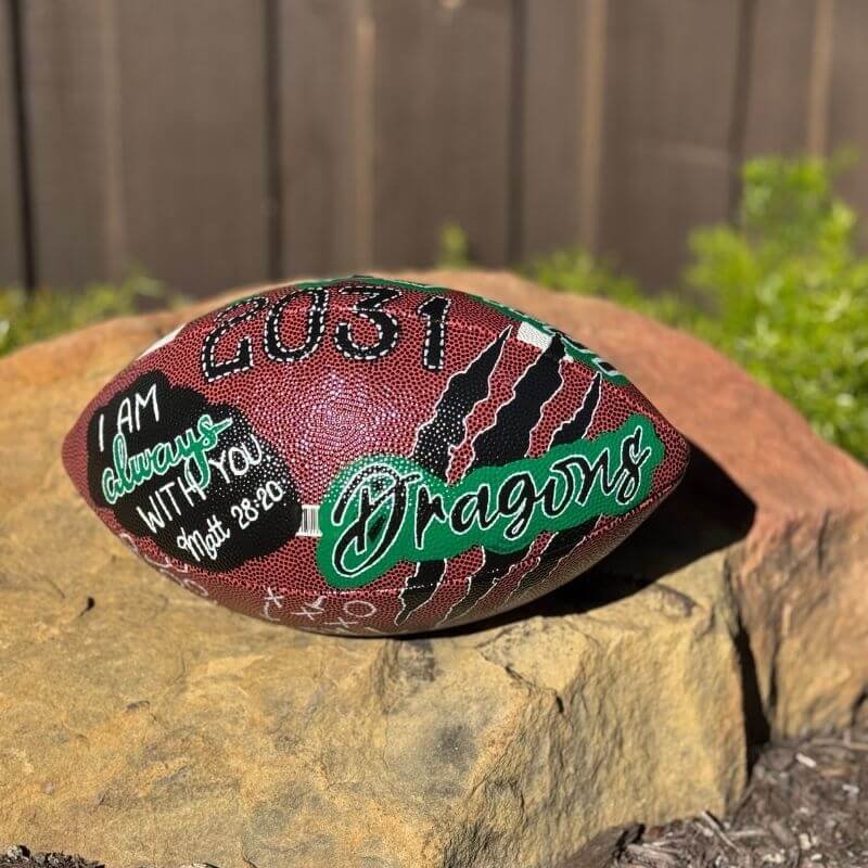 Decorated football with "I am always with you! Matt 28:20", "2031", and "Dragons" text placed on a rock.