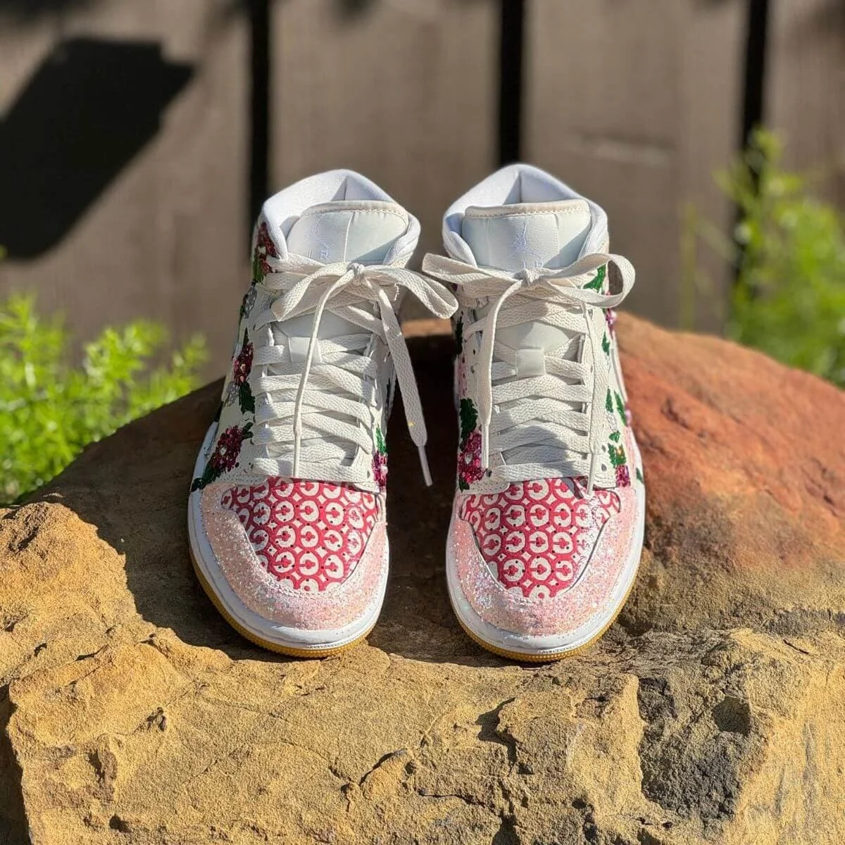 Pair of sneakers with floral and geometric patterns on a rocky surface, with a wooden fence and greenery in the background.