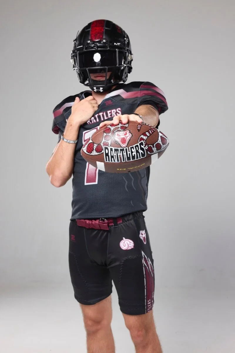 High school football player in a black and red Rattlers uniform holding a custom-painted football featuring the Rattlers mascot.