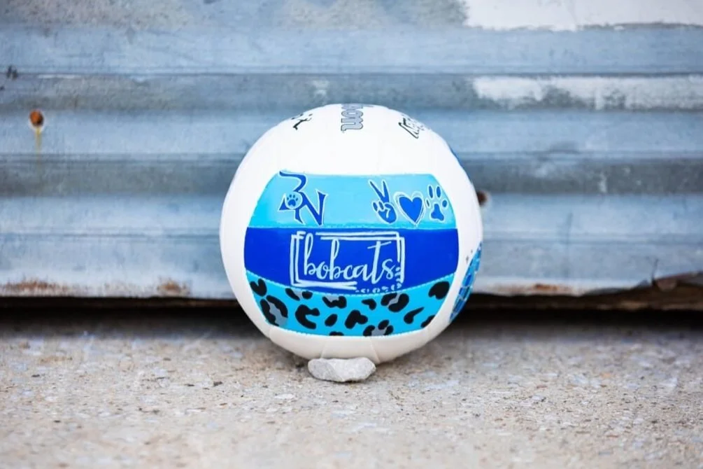 Custom white volleyball with blue and black accents, featuring the words 'Bobcats' along with peace signs and heart icons, resting against a metal background.