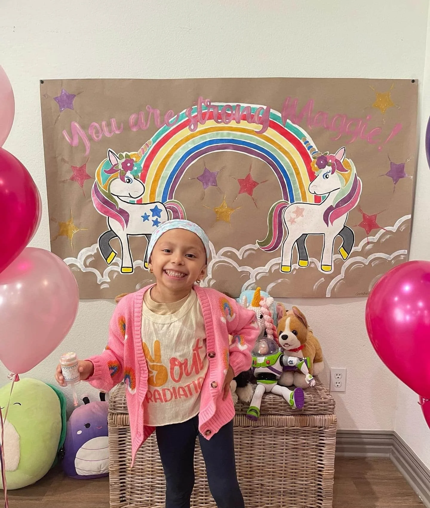 A smiling young girl standing in front of a colorful unicorn and rainbow backdrop with the message "You are magical!" She is holding a small bottle and wearing a pink cardigan with rainbows.