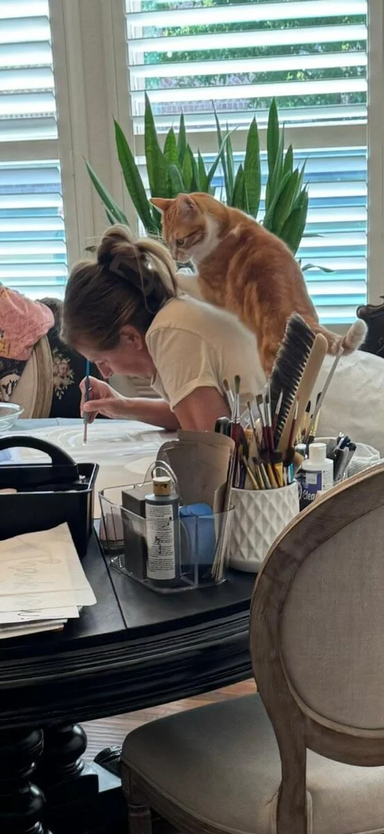 artist leaning over table hand painting a banner with orange cat supervising