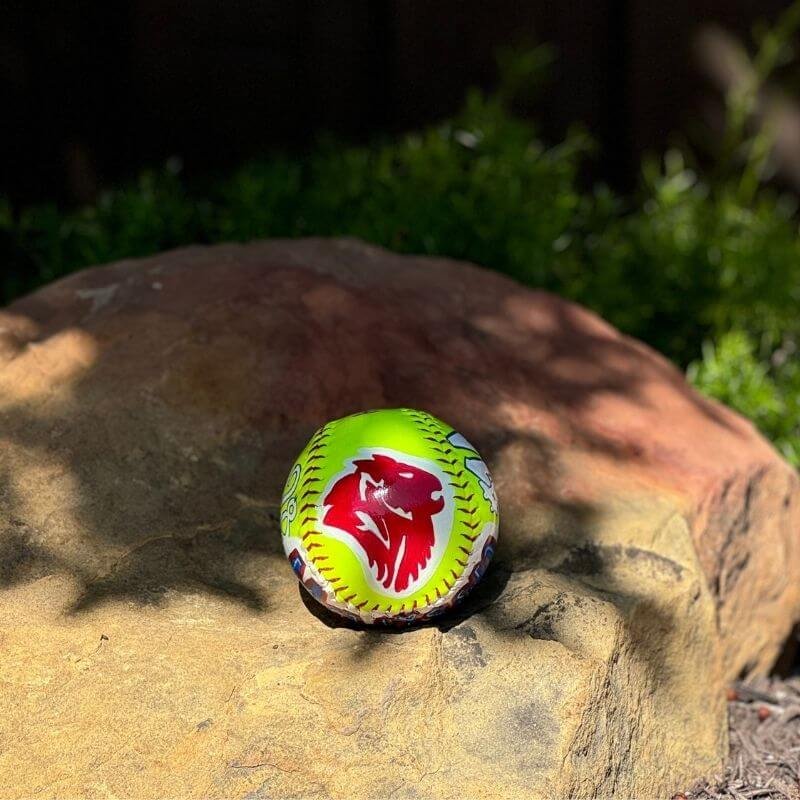 A neon yellow softball with a red logo on a rock outdoors.