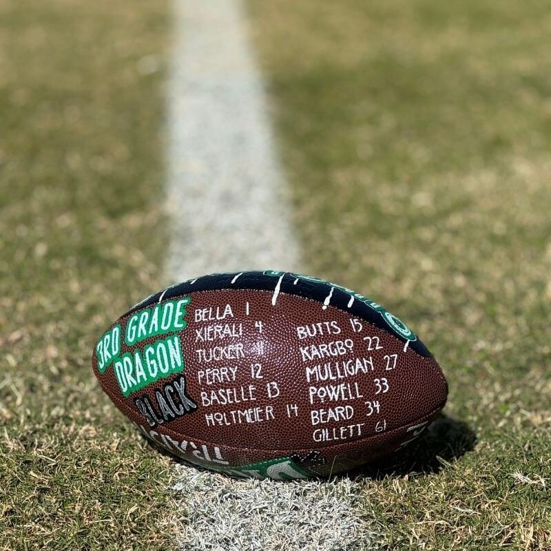 A football on a grass field featuring the text "3rd Grade Dragon Black" and a list of names and numbers written on it. The ball is placed on the white sideline.