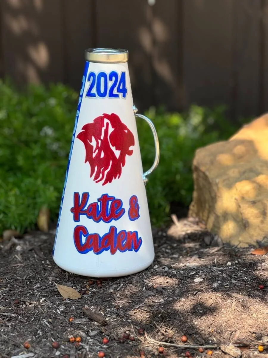 A white megaphone with "2024" and "Kate & Dalen" written on it, sitting on a rock with plants.