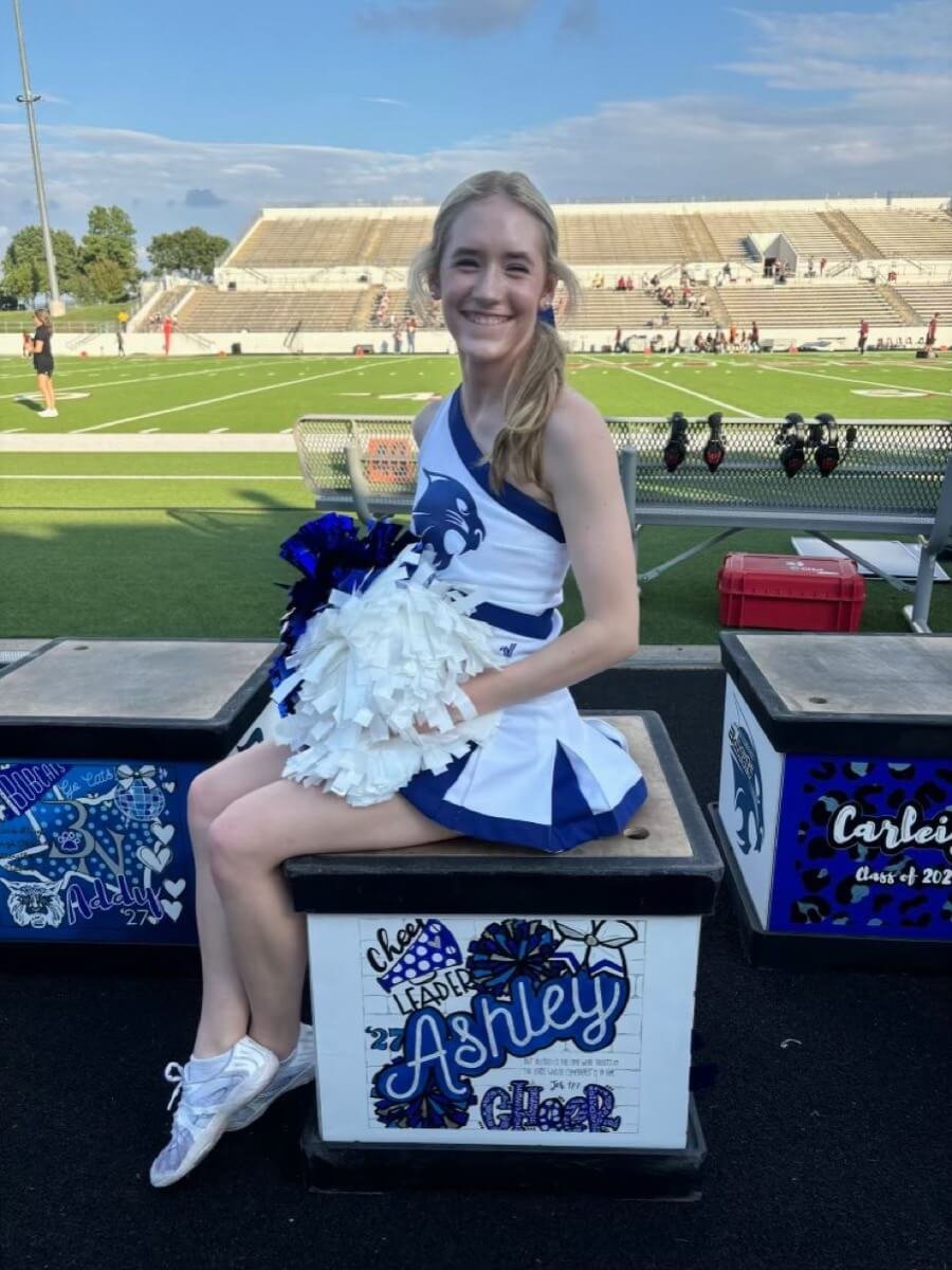 Cheerleader Ashley posing with custom-painted cheer box, smiling on the field.