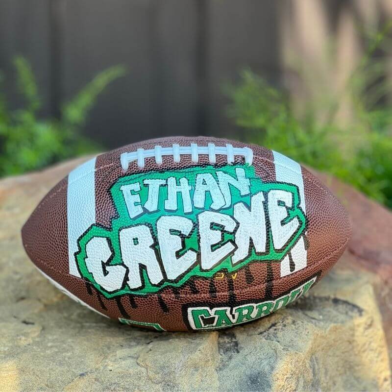 A football with graffiti-style text displaying the name "Ethan Greene." The ball is placed on a rock outside, with grass visible in the background.