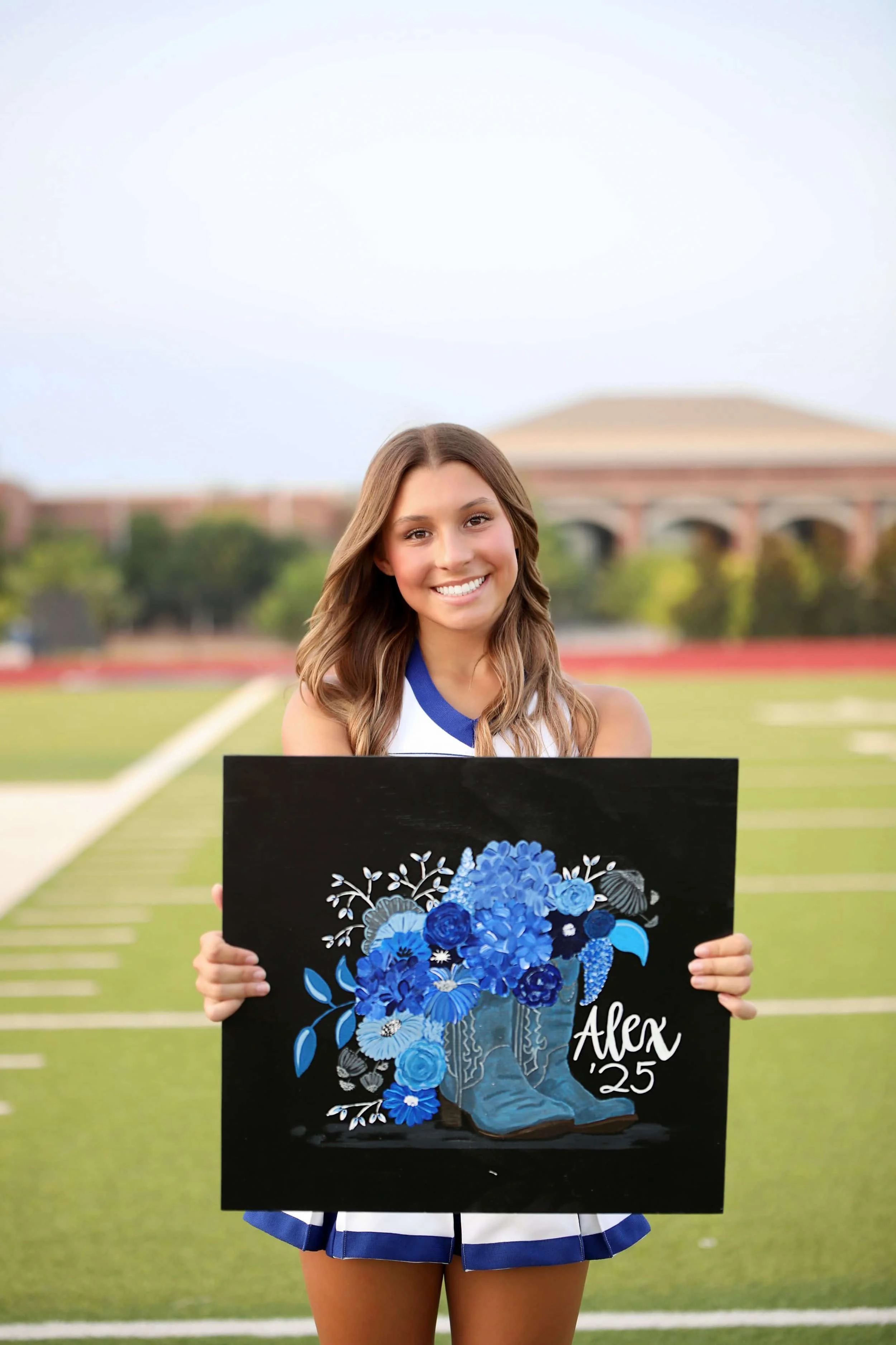 A young woman in a cheerleading uniform holding an art piece with blue flowers and a pair of blue cowboy boots, on a football field with a school building in the background.