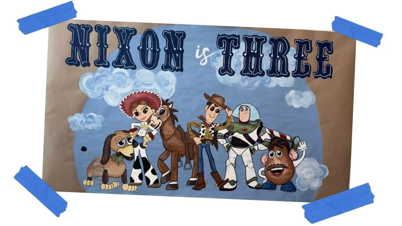Custom hand-painted "Nixon is Three" birthday banner featuring Toy Story characters Woody, Jessie, Bullseye, Buzz Lightyear, and Mr. Potato Head on a blue sky background, taped up with blue painter's tape.