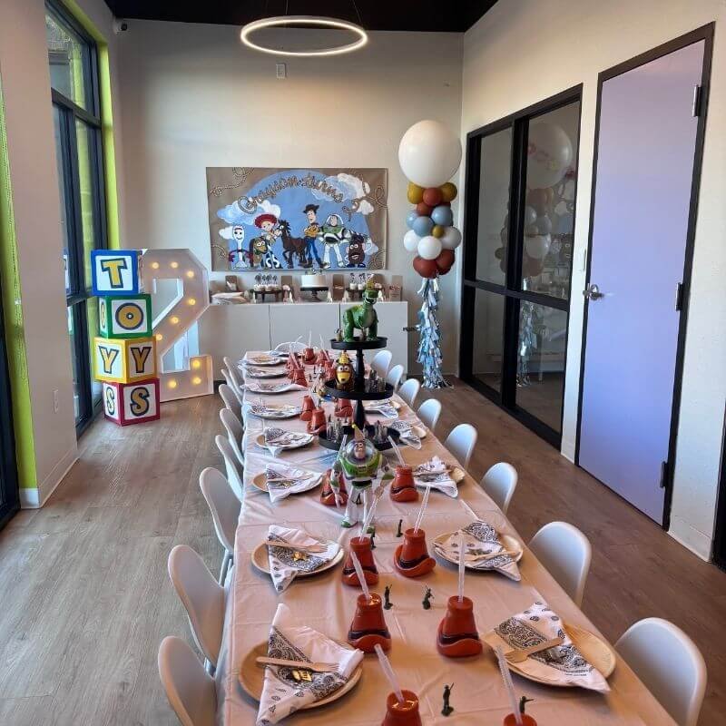 A Toy Story-themed birthday party setup with a long table, themed decorations, and character figurines. A decorated backdrop features Toy Story characters. Marquee letters spelling 'TOYS' and a large number '2' are displayed. There's a balloon arrang