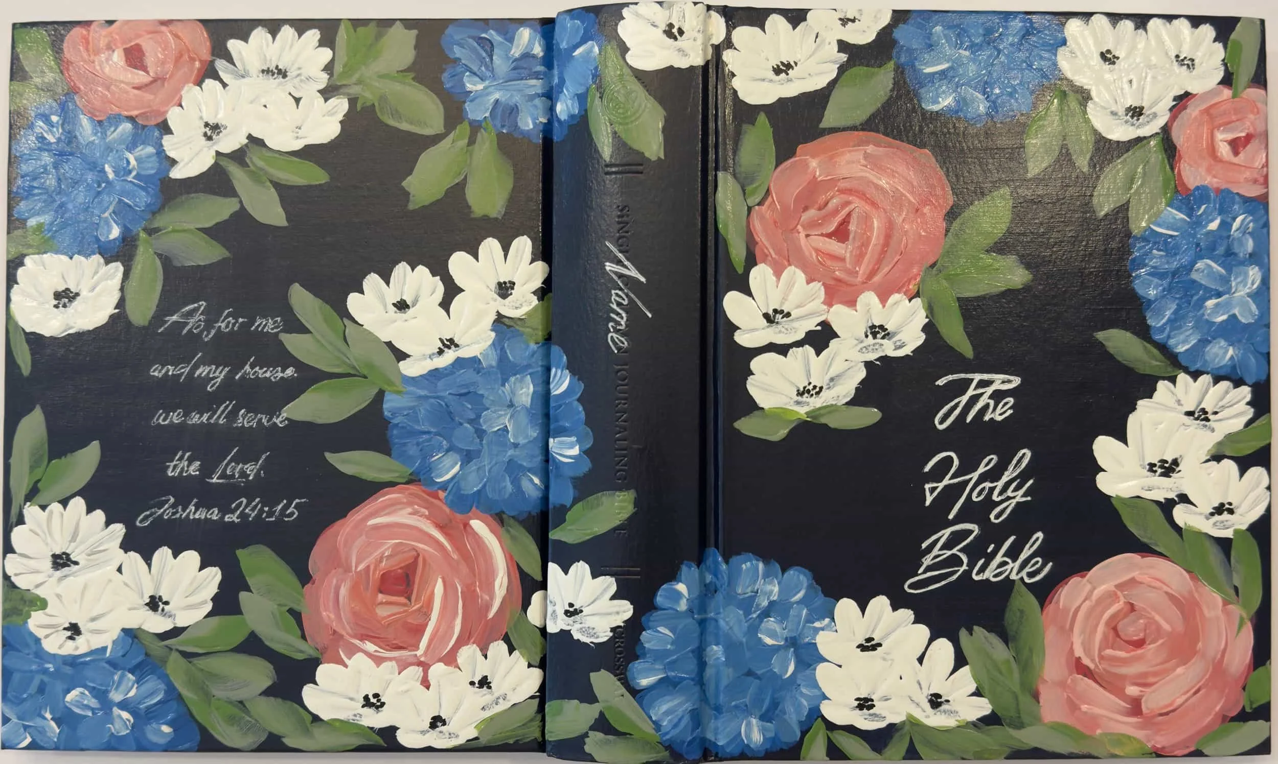 An illustrated cover of the Holy Bible with a floral design featuring pink roses, white daisies, blue hydrangeas, and green leaves on a black background. There is a handwritten quote from Joshua 24:15 on the left side and the title 'The Holy Bible' o