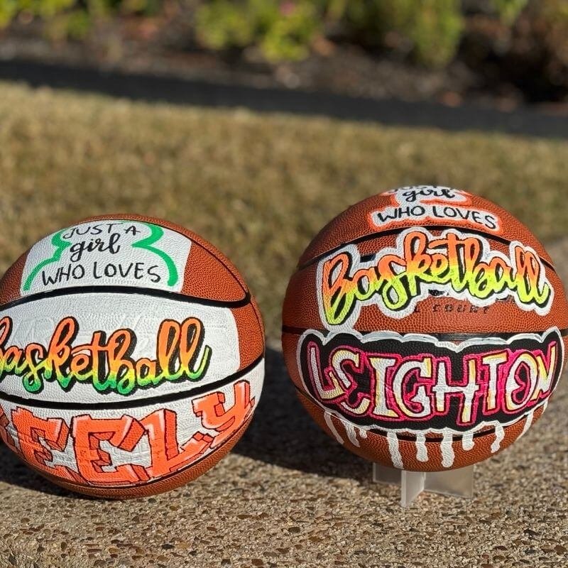 Two customized basketballs with colorful, hand-painted designs and names. The left ball says "Just a girl who loves basketball" and includes the name FELY. The right ball has "girl who loves basketball" and the name LEIGHTON written on it.