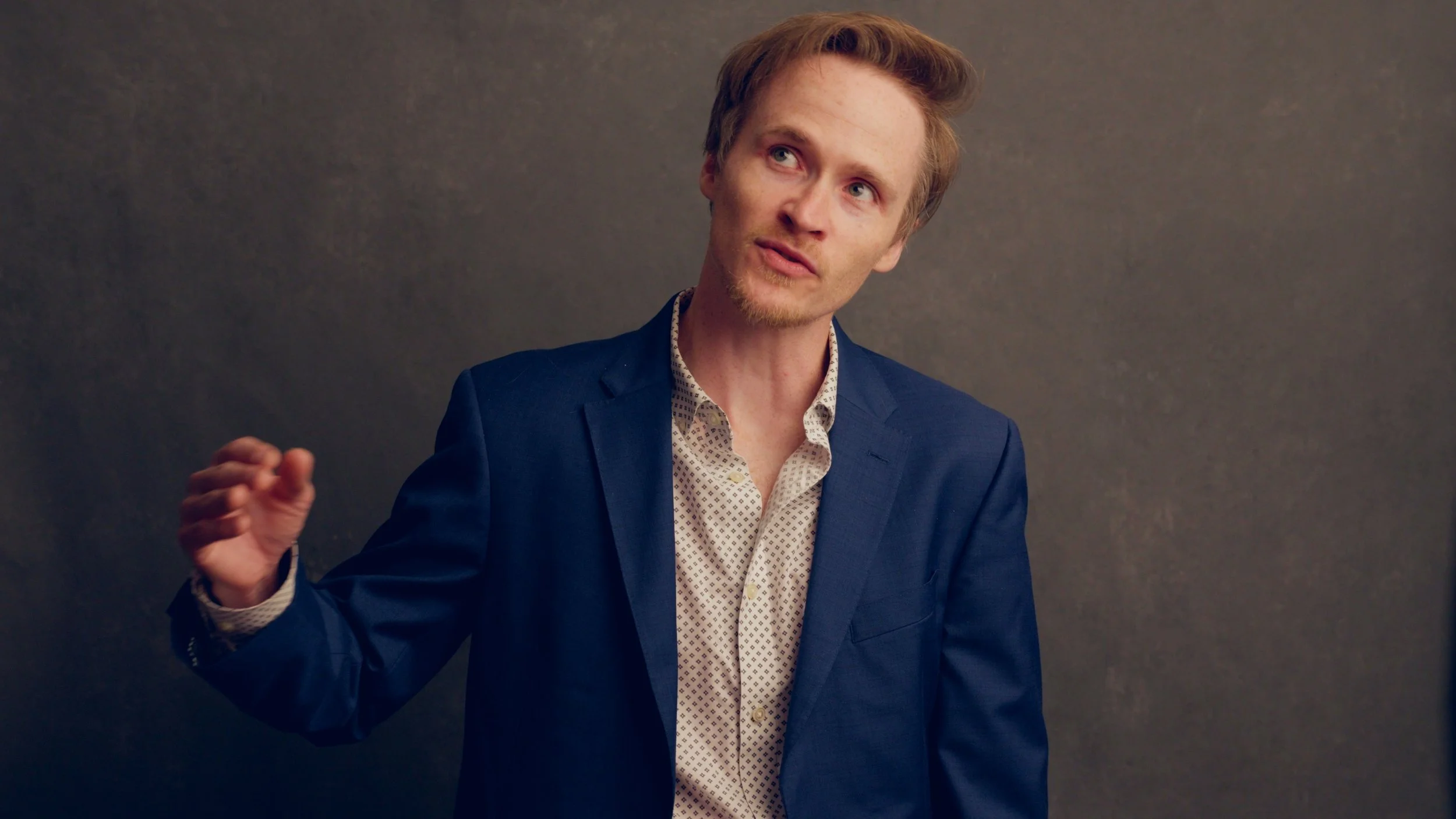 A man with red hair and a goatee, wearing a navy blazer and patterned shirt, speaking or explaining something against a plain gray wall.