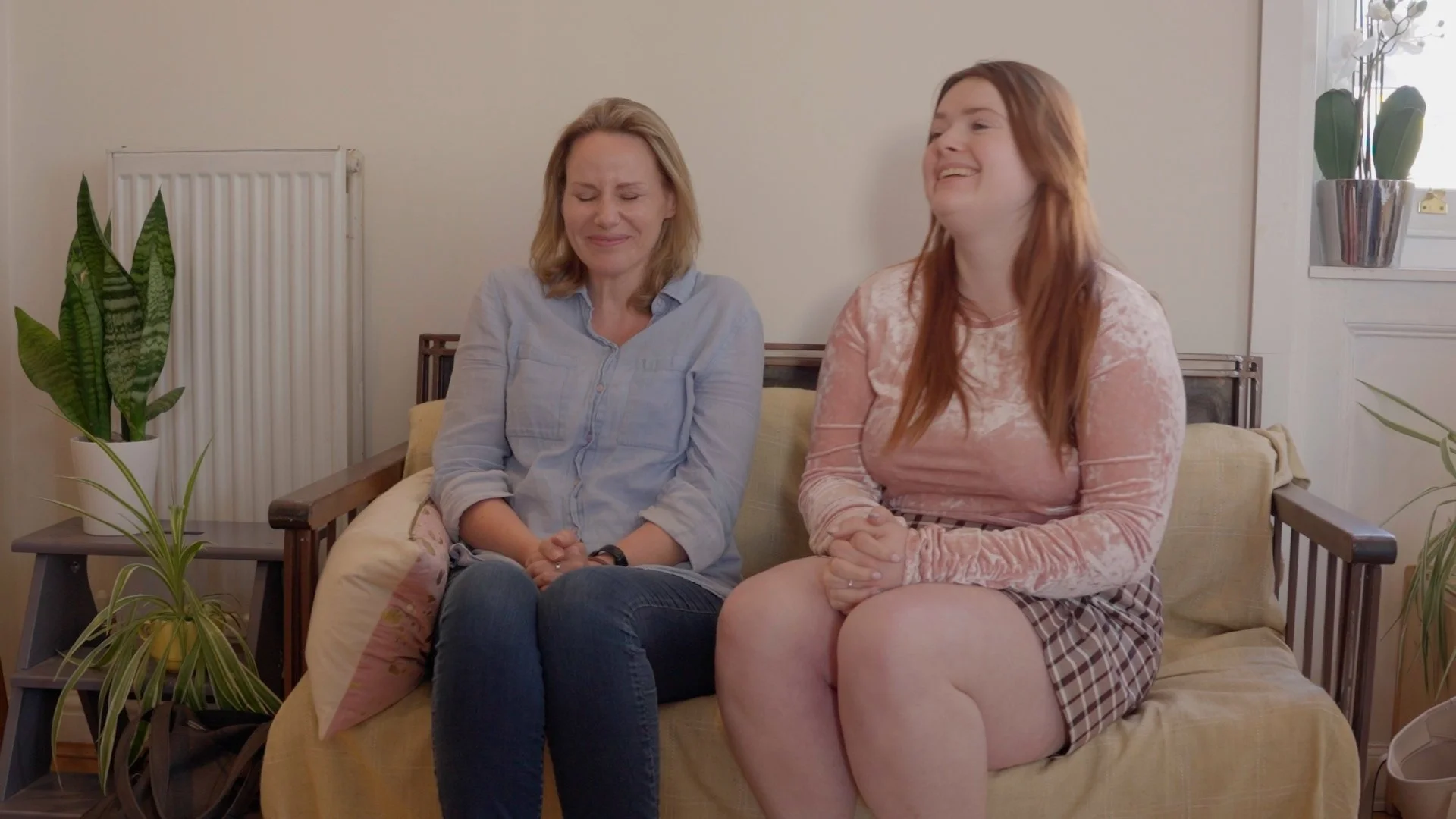 Two women sitting on a beige couch, one with eyes closed and the other smiling with eyes closed, in a brightly lit room with plants.