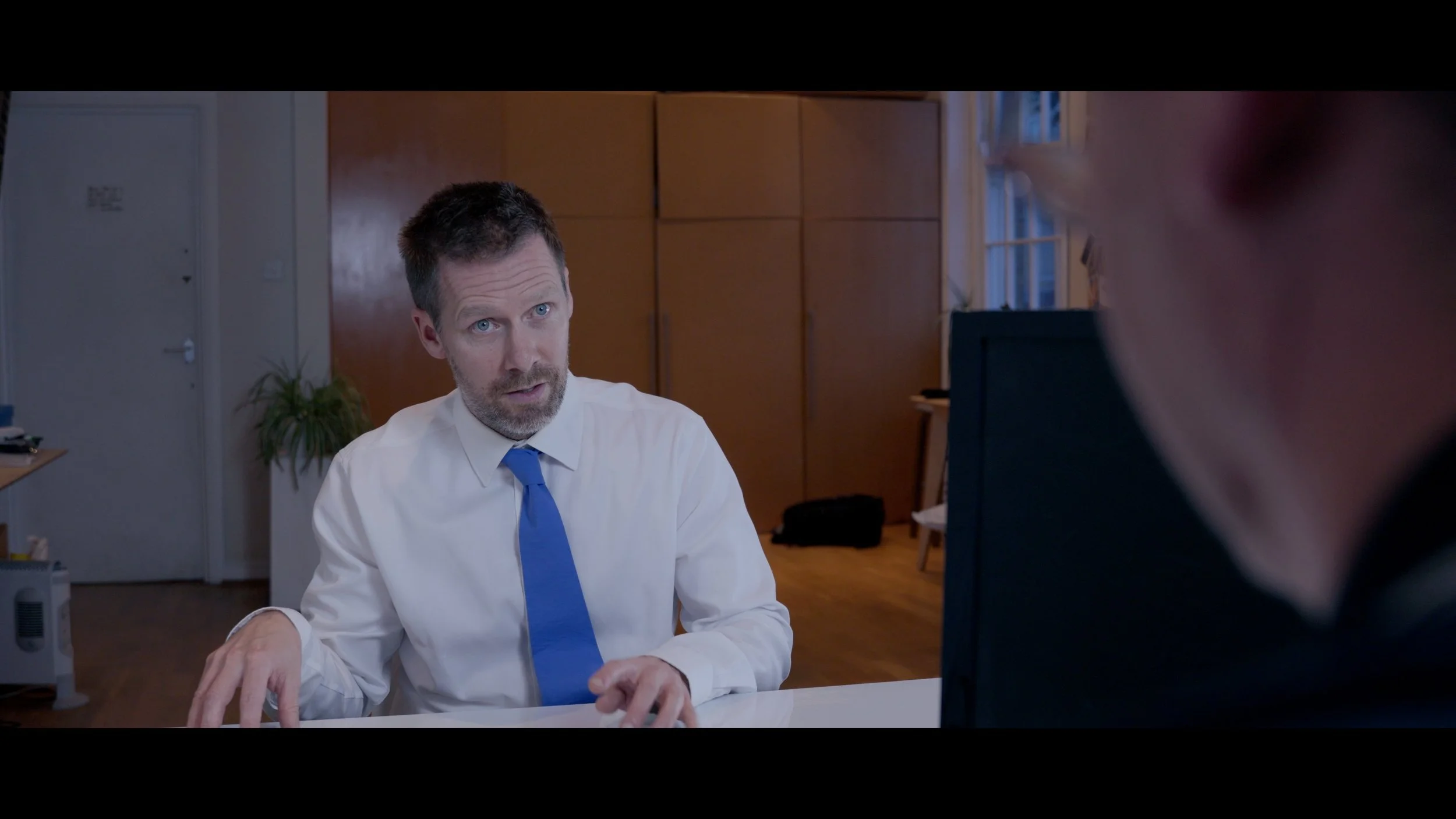 Still image taken from an acting scene in which a male actor is sitting across from another actor at a desk